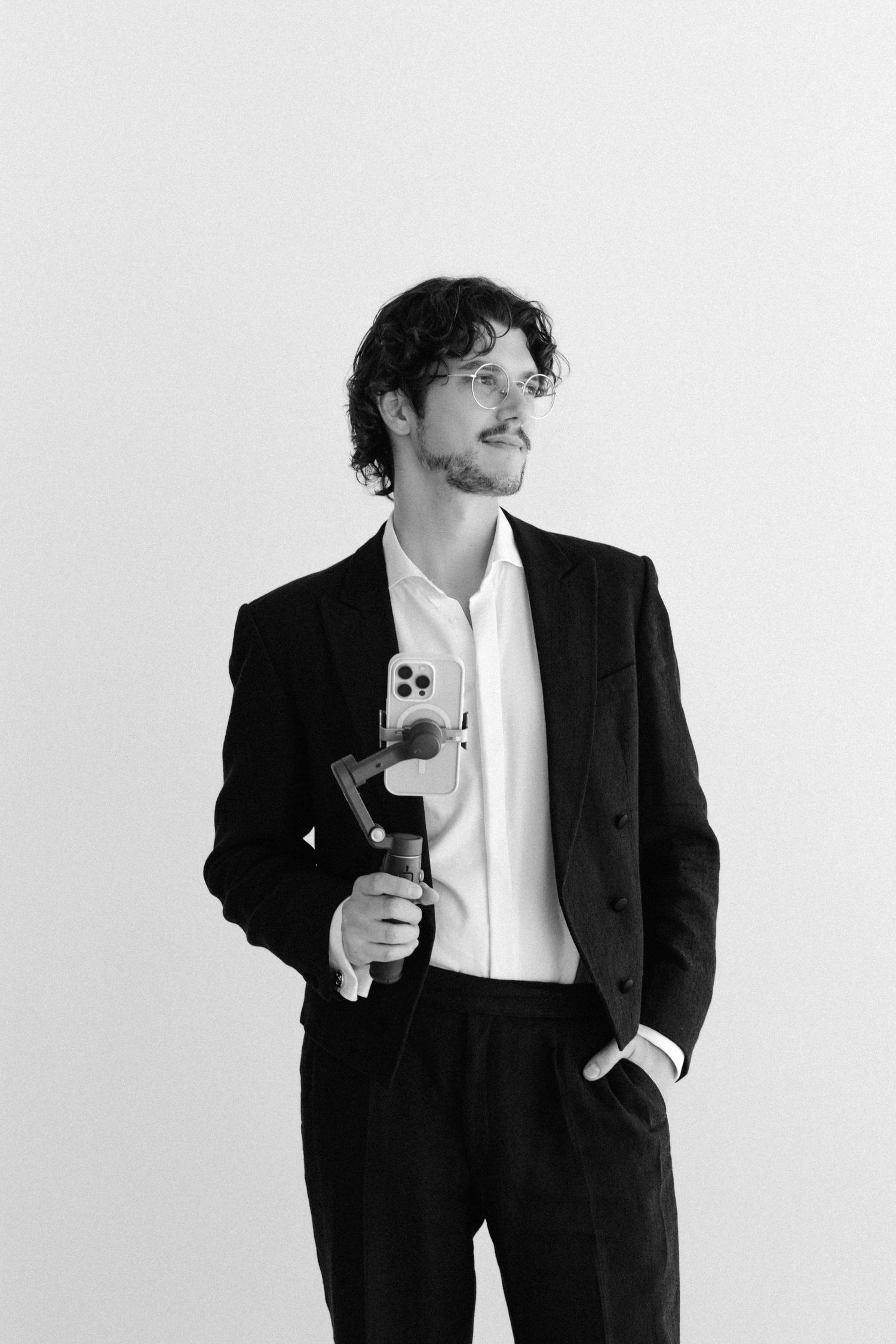 Black and white portrait of a young man in a suit holding a smartphone gimbal stabilizer against a white background.