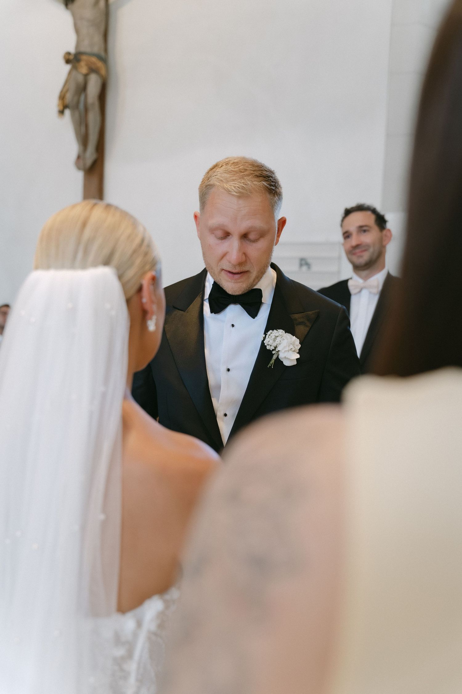 Groom in a black tuxedo with bow tie and white boutonniere speaks during a church wedding ceremony as a veiled bride faces him.