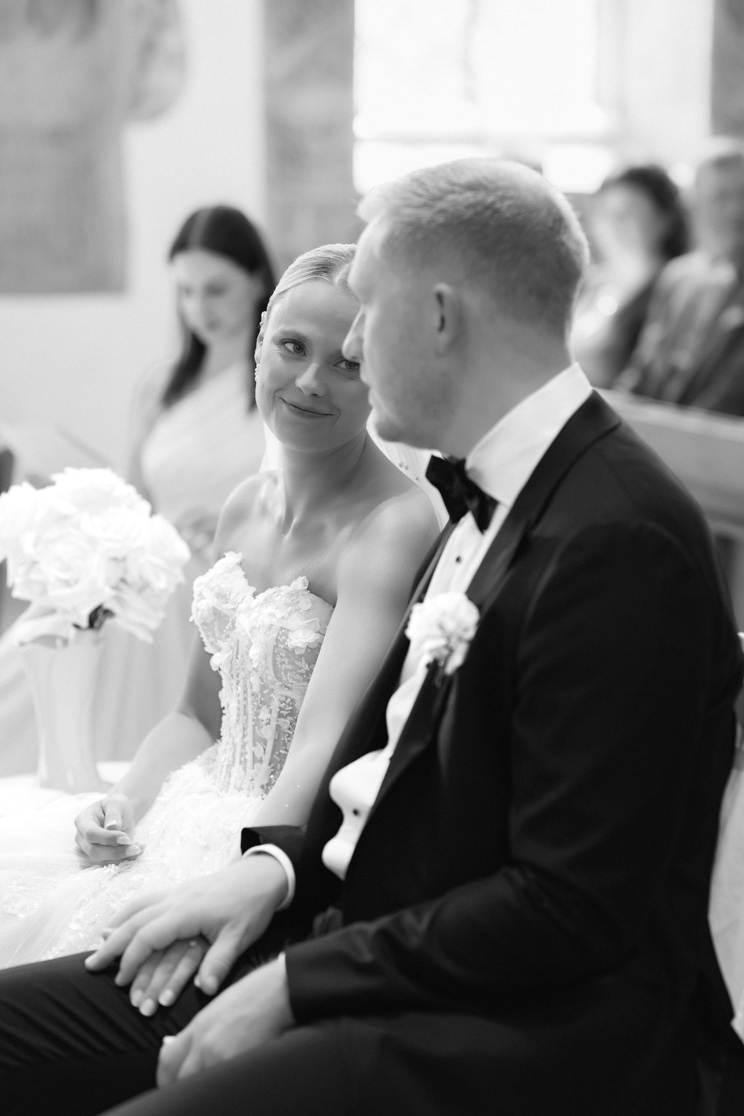 Black and white photo of a bride in a lace gown smiling at the groom in a tuxedo as they sit together during a church wedding ceremony.