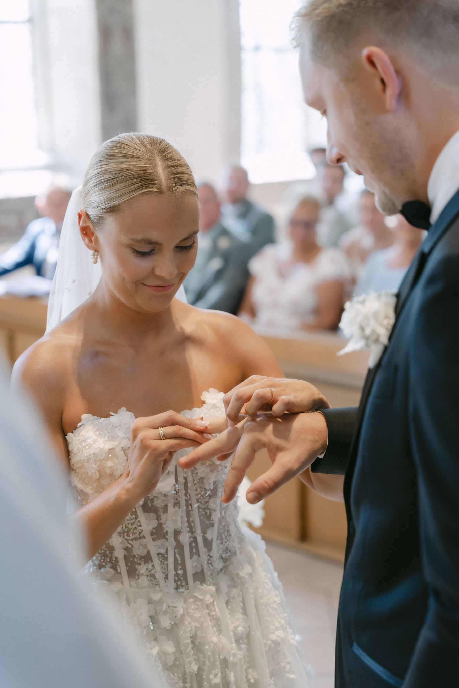A smiling bride in a lace strapless gown places a ring on the groom's finger during a wedding ceremony as guests watch.