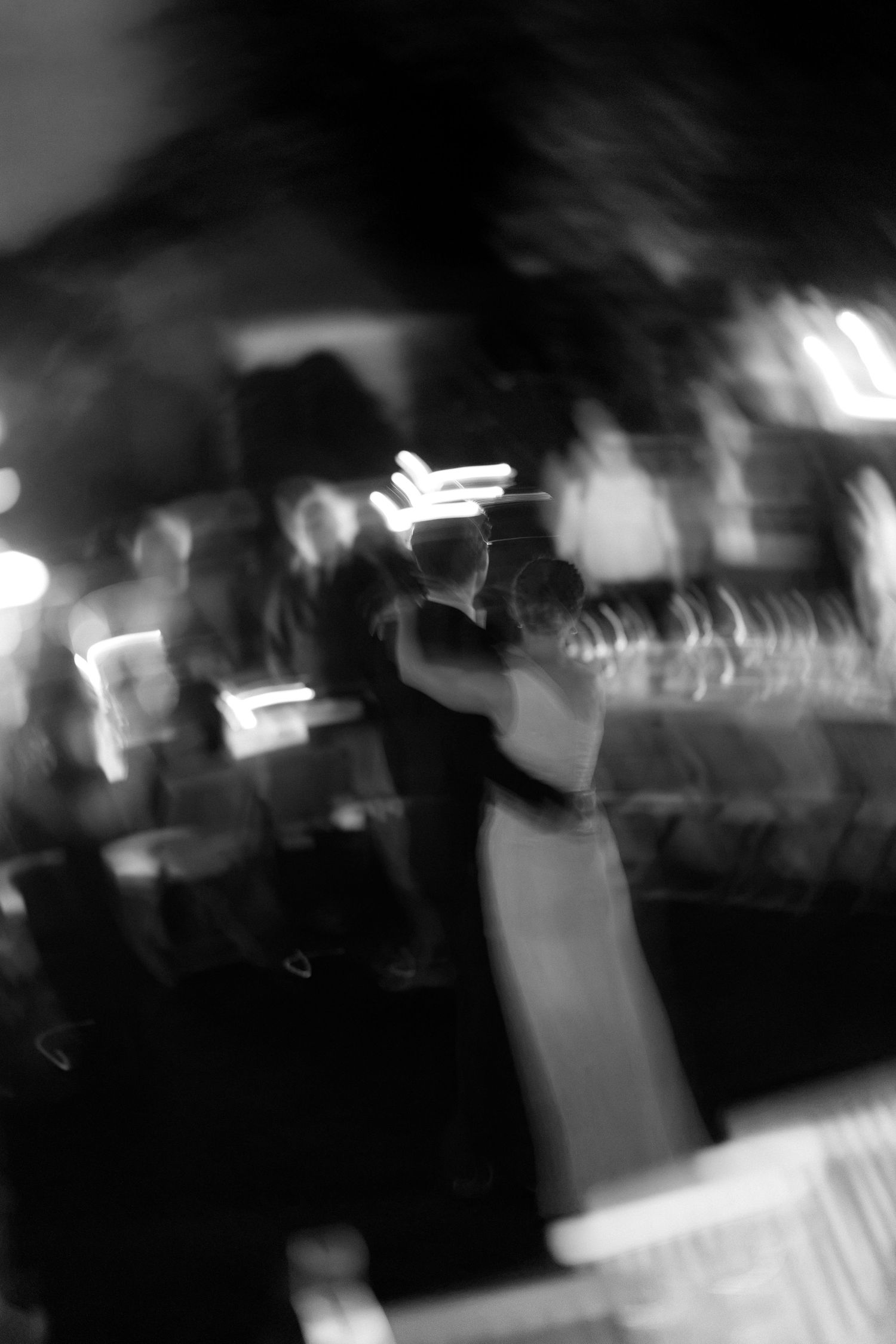 Blurred black and white photo of a couple slow dancing at an evening reception with glowing lights and guests in the background.