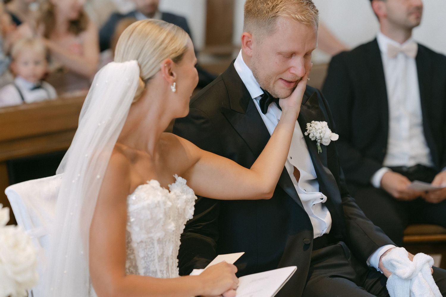 A bride in a lace gown and veil gently wipes a tear from the emotional groom's cheek during a wedding ceremony as guests look on.