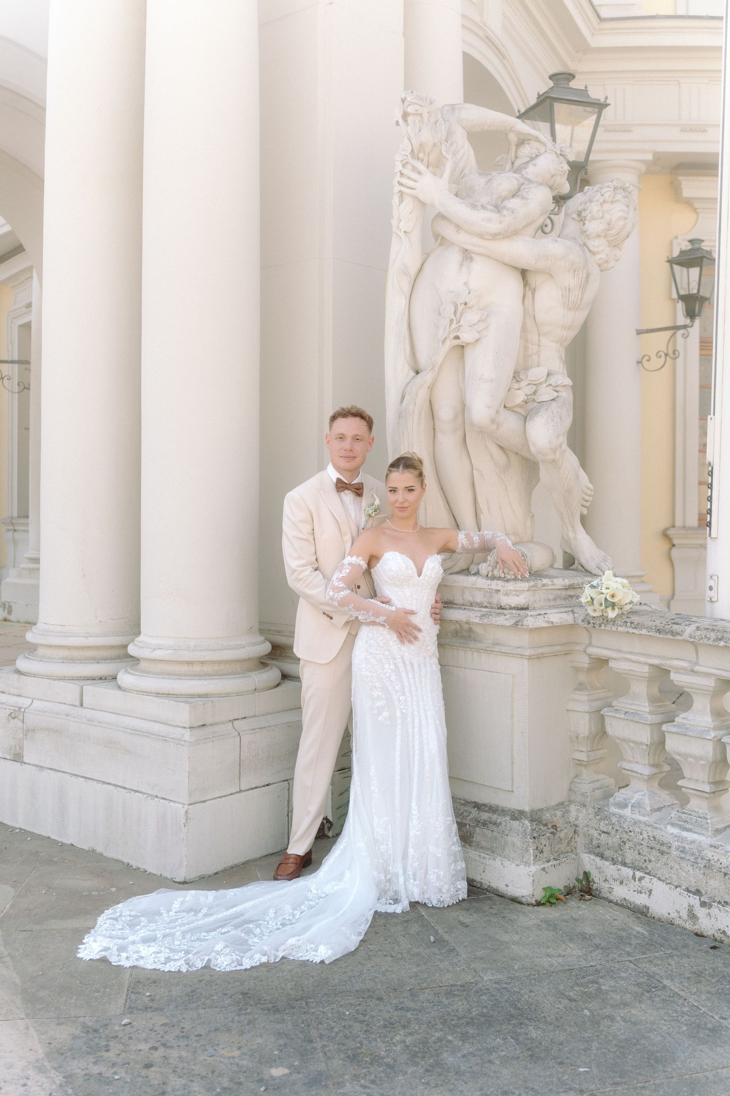 Elegant wedding couple poses against ornate white marble columns and statues in a classical architectural setting. Schloss Ludwigsburg