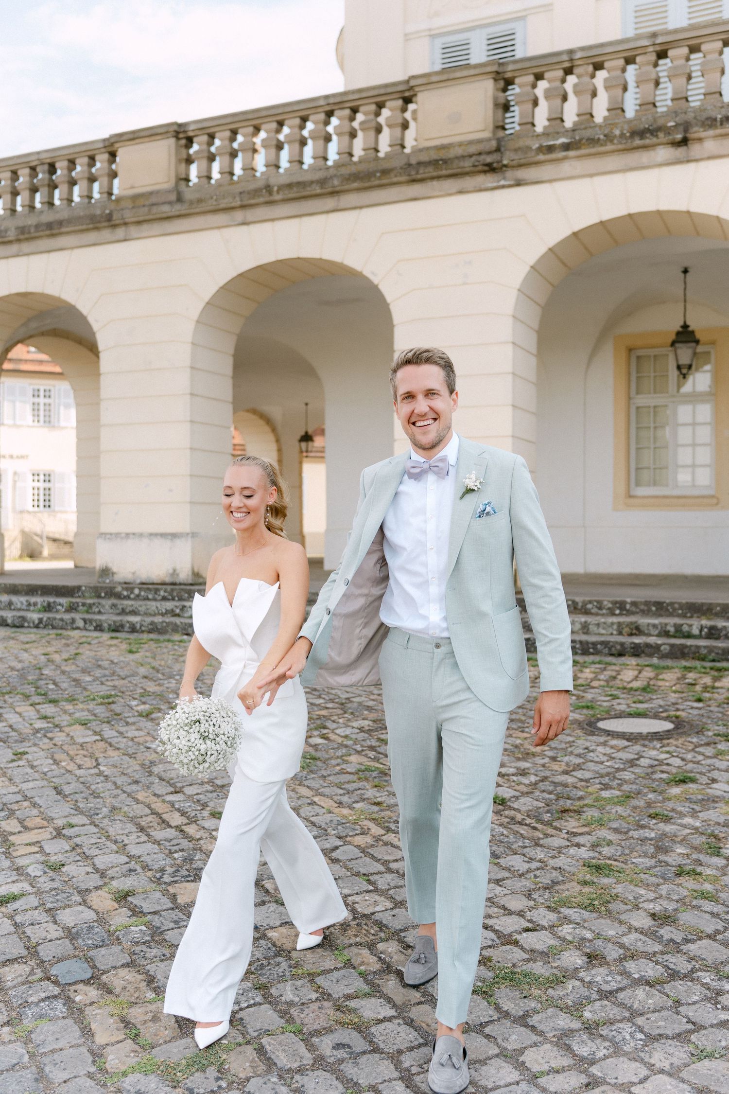 A couple in wedding attire shares playful moments under an elegant stone archway with cobblestone ground. Schloss Solitude