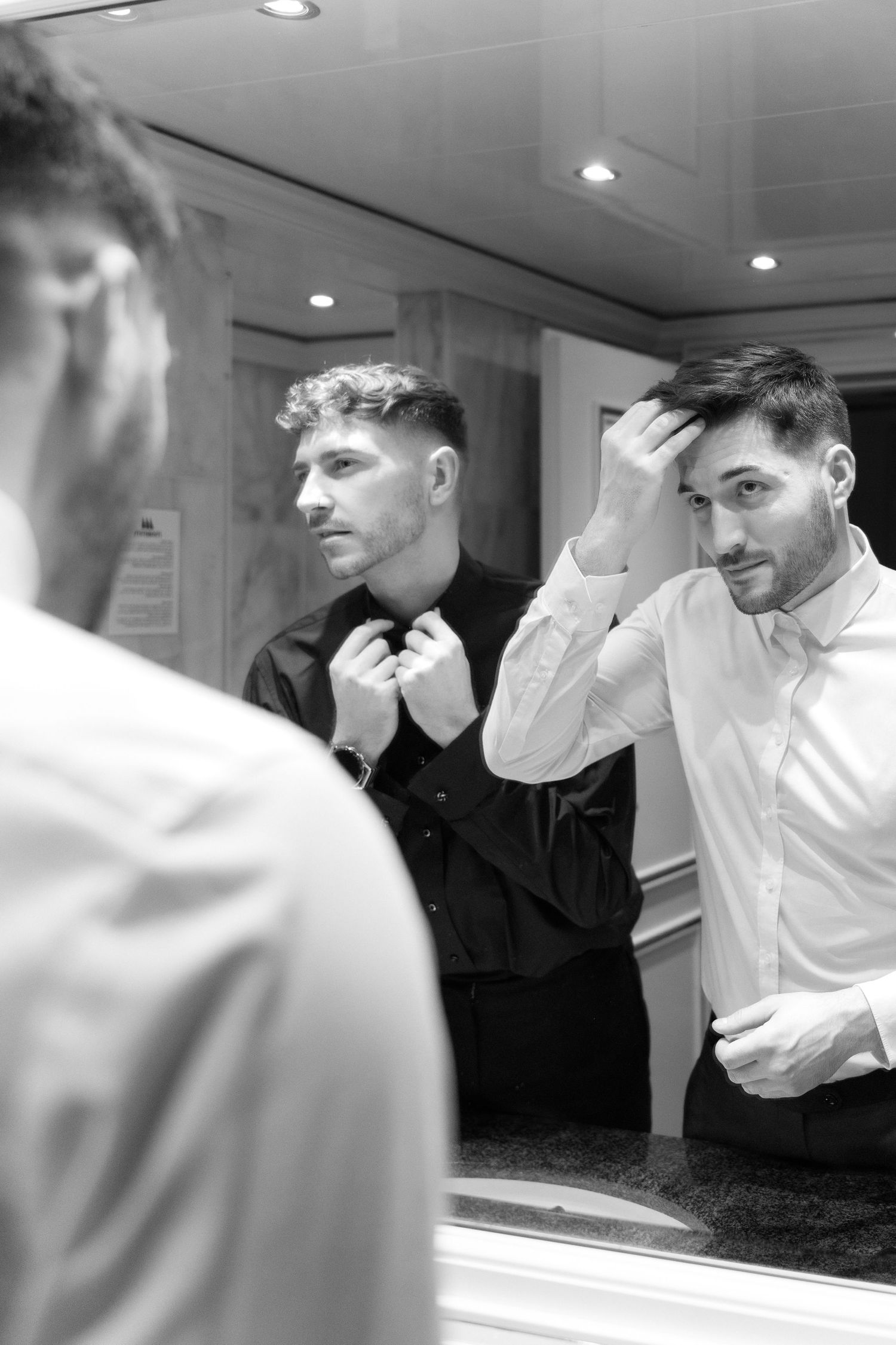 Black and white reflection in mirror showing well-dressed men adjusting their formal attire in an indoor setting.