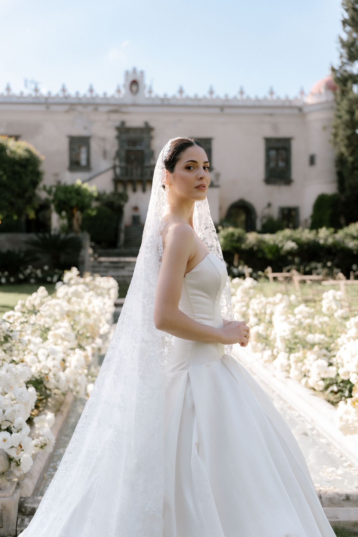 Bride in a white ball gown and lace veil poses in a garden with white flowers and an elegant mansion backdrop.