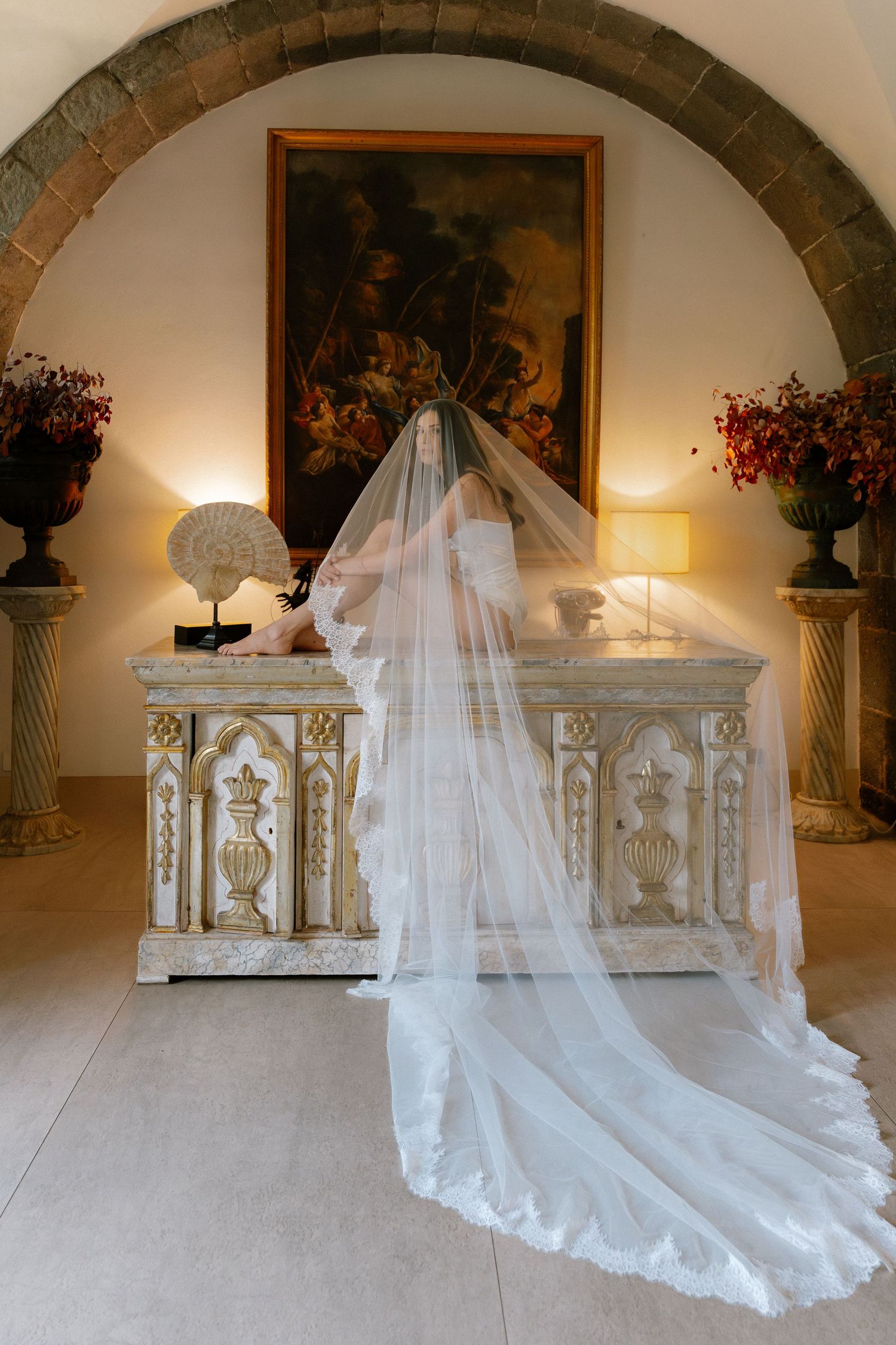 Bride in lace wedding gown and long cathedral veil stands before ornate antique dresser under stone arch in elegant venue.