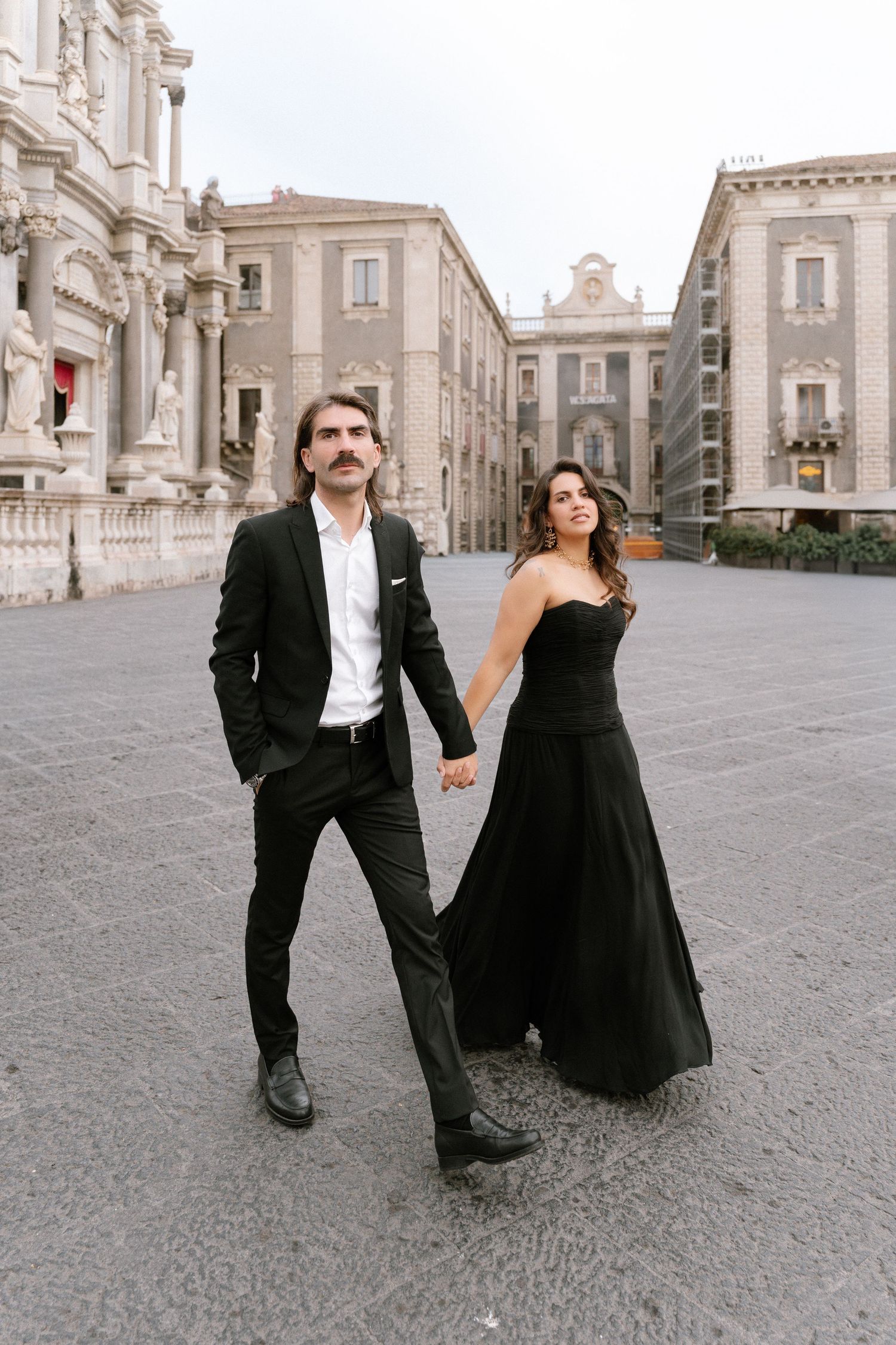 Elegant couple in matching black formal attire holding hands in a European plaza with historic baroque architecture.