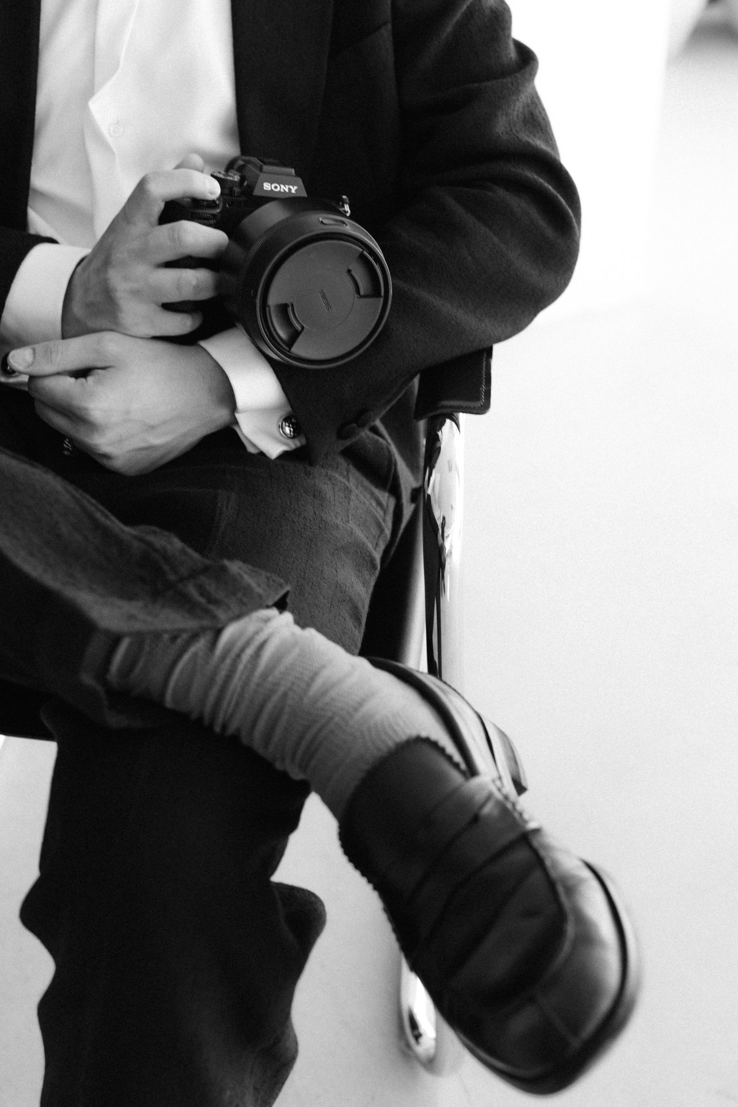 Black and white photo of a person in a suit holding a Sony camera while seated, wearing dress shoes.