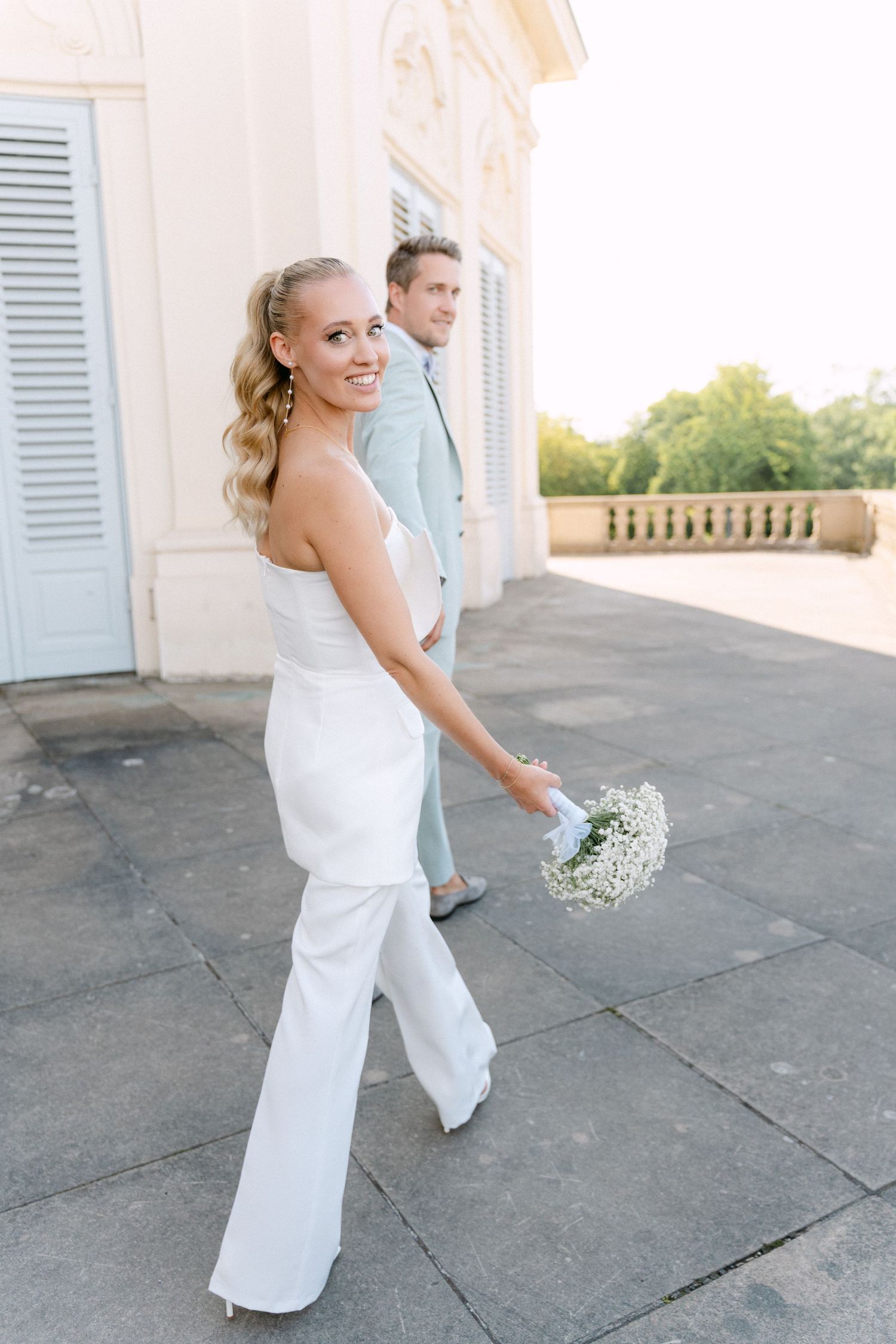 Modern wedding couple in white and light blue attire pose on an elegant stone terrace with a delicate floral bouquet. Schloss Solitude