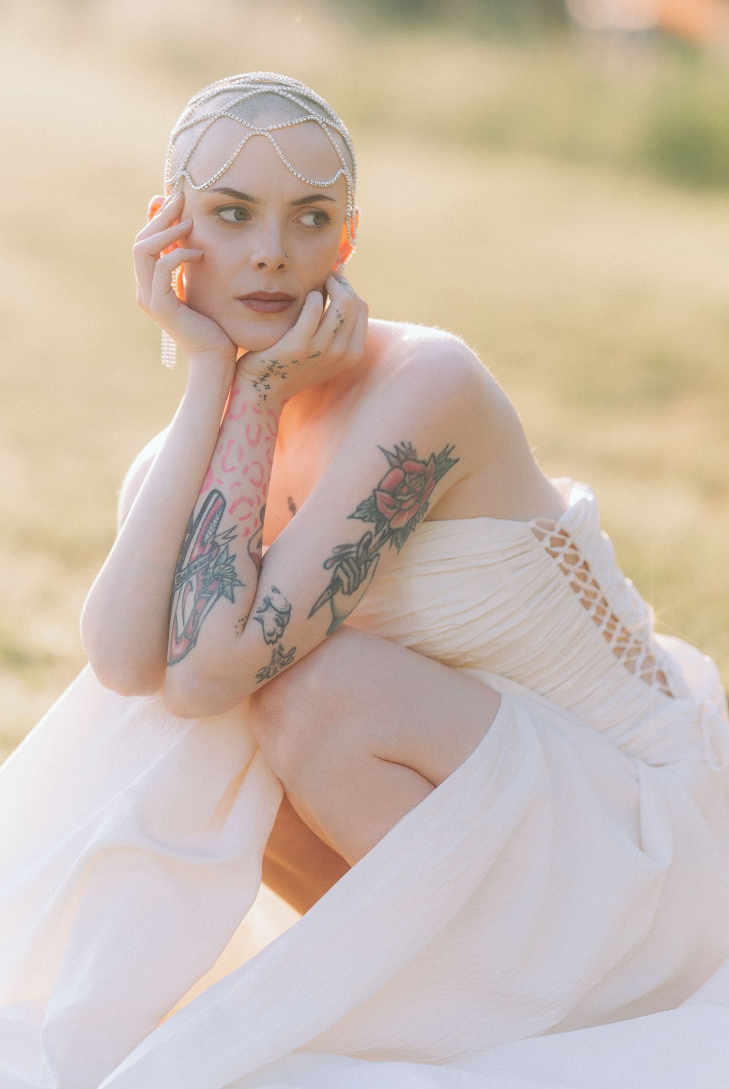 A person in a flowing white dress poses outdoors at sunset, showing artistic tattoos on their arms in dreamy lighting.
