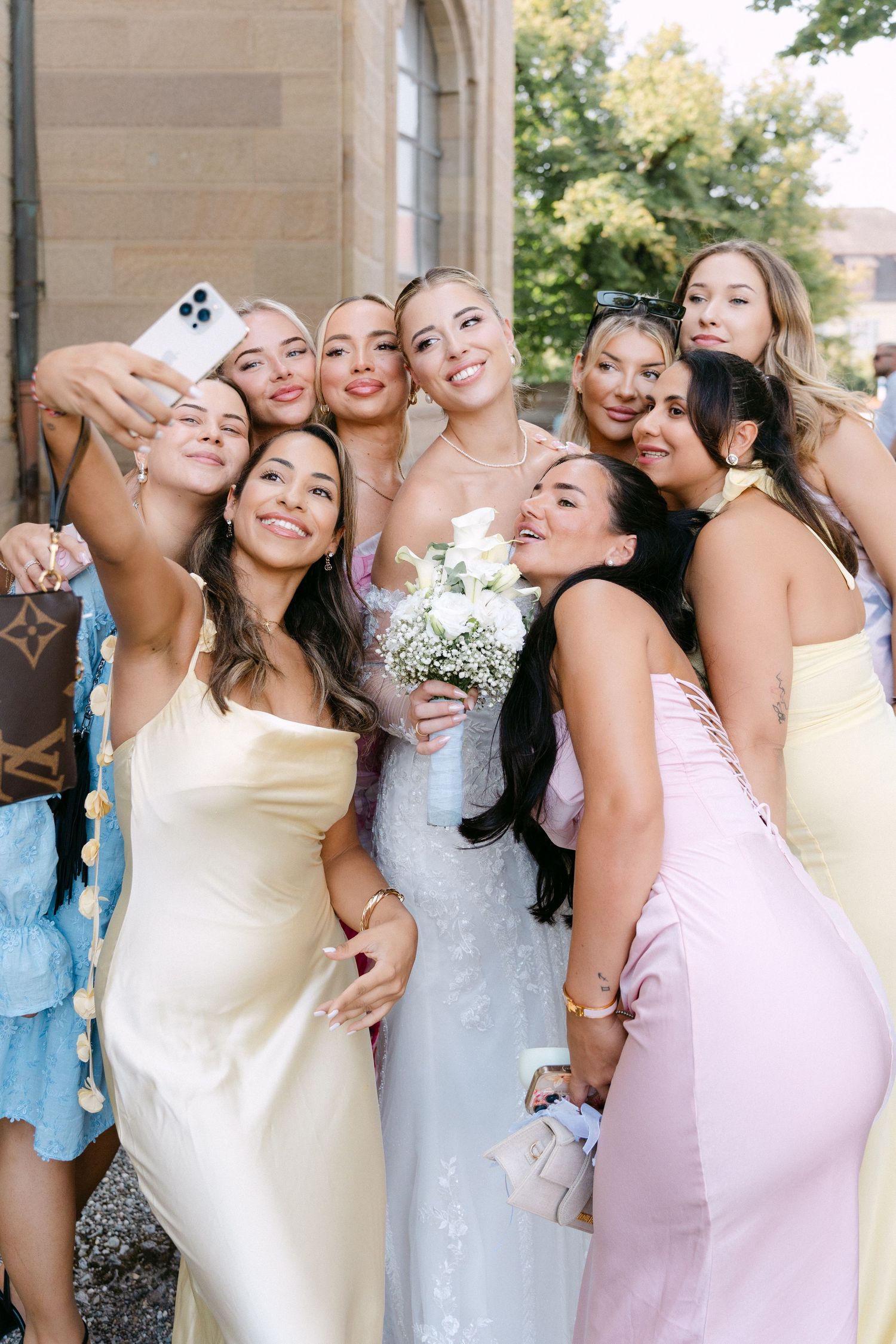Bridesmaids in pastel dresses and white gowns pose together playfully at an outdoor wedding celebration.