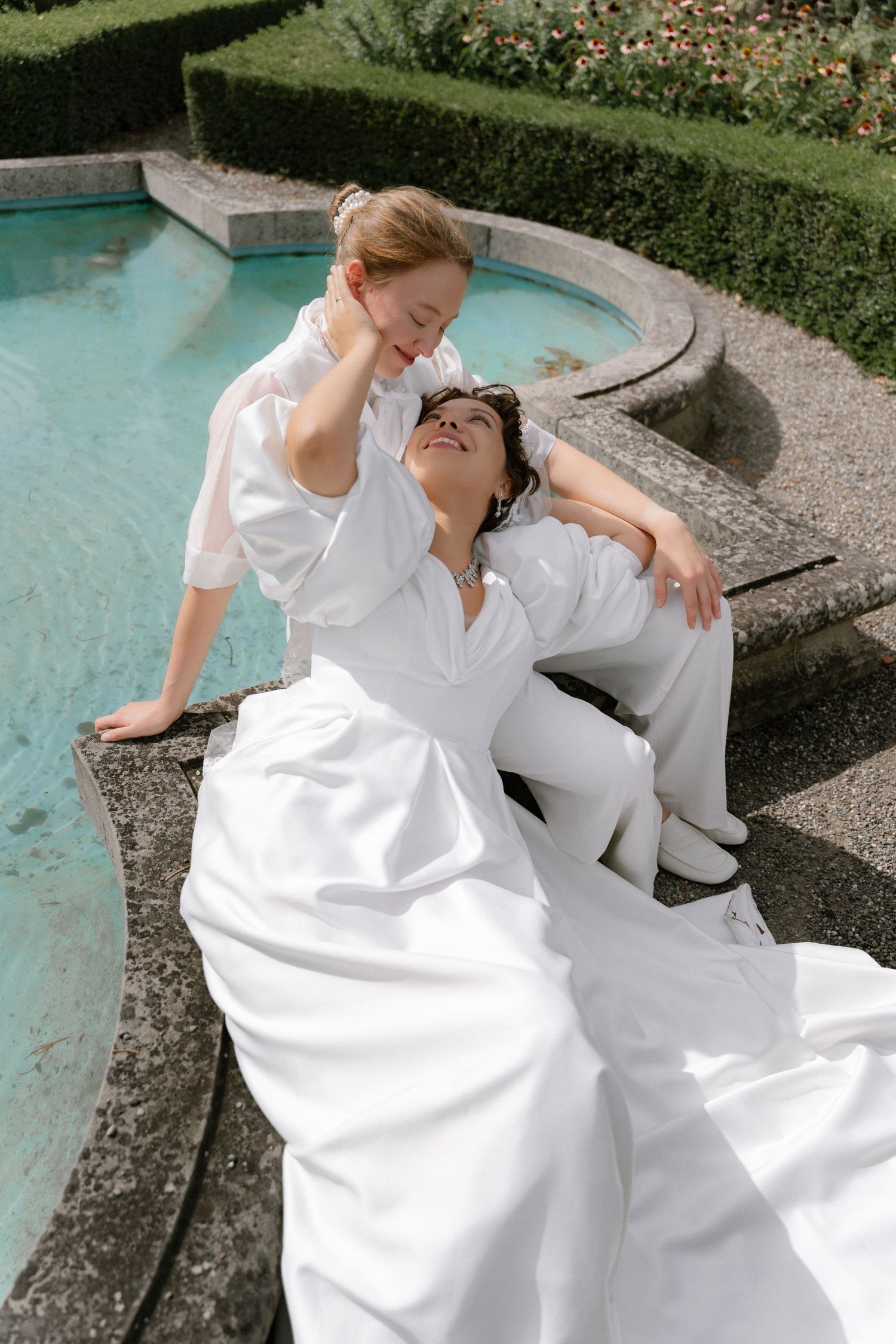 Two brides in white attire sit elegantly by a garden fountain, sharing a tender moment outdoors.