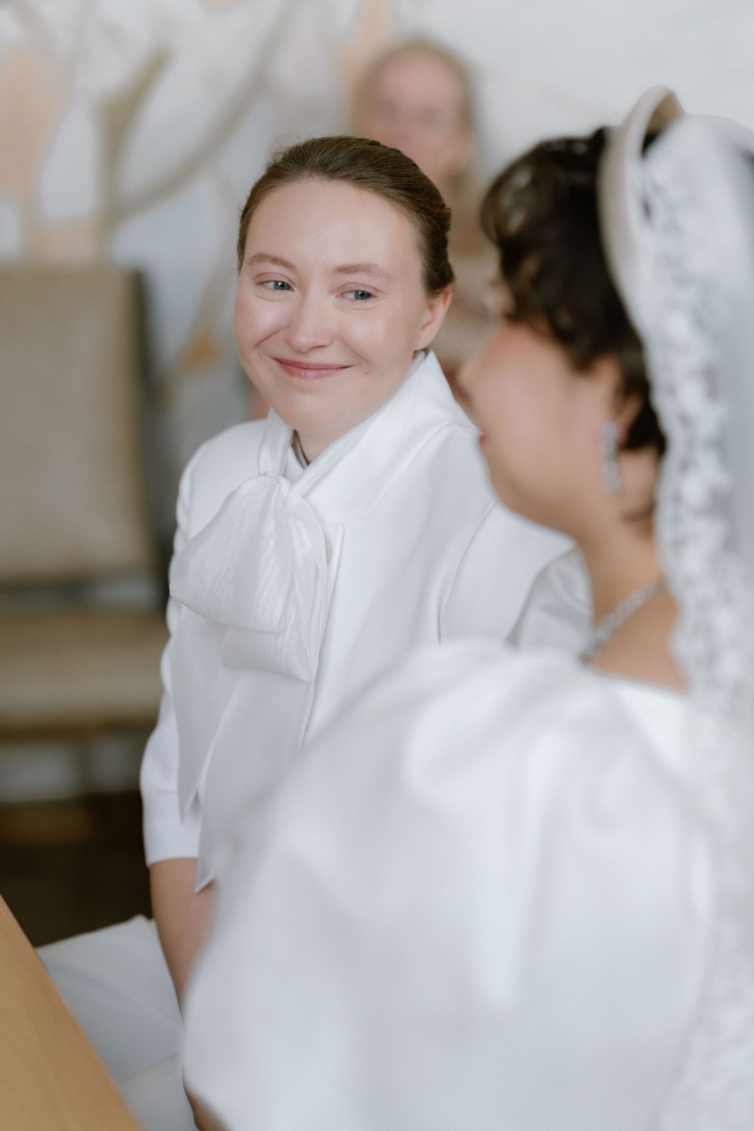A smiling woman in a white blouse looks at a bride wearing a veil during a wedding ceremony.