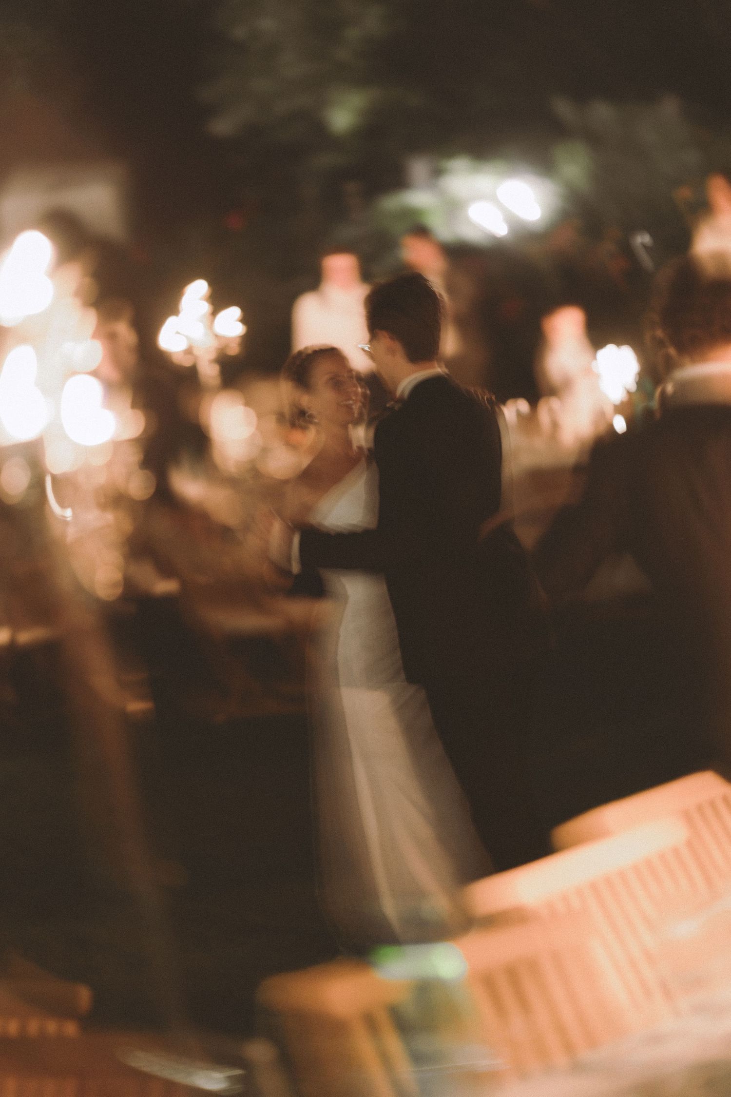 Blurred bride and groom dancing together at an evening outdoor wedding reception with glowing string lights and wooden chairs.