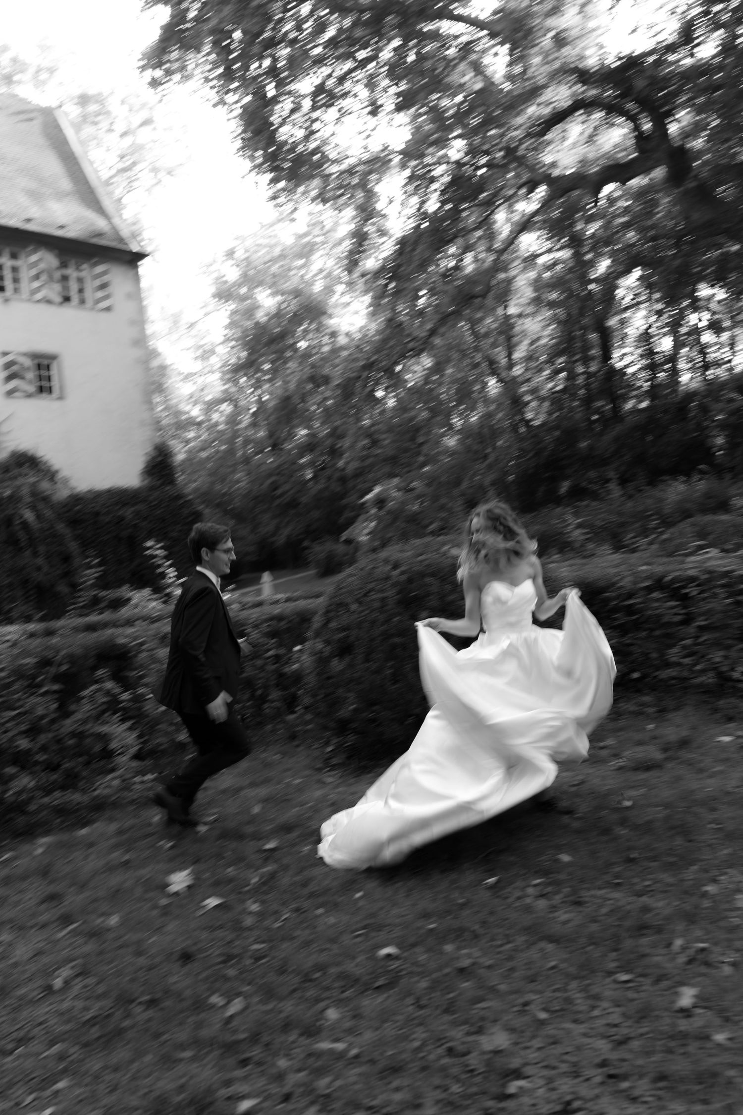 Black and white photo of a bride in a flowing gown twirling on a grassy lawn as a groom in a suit walks toward her near a historic house.