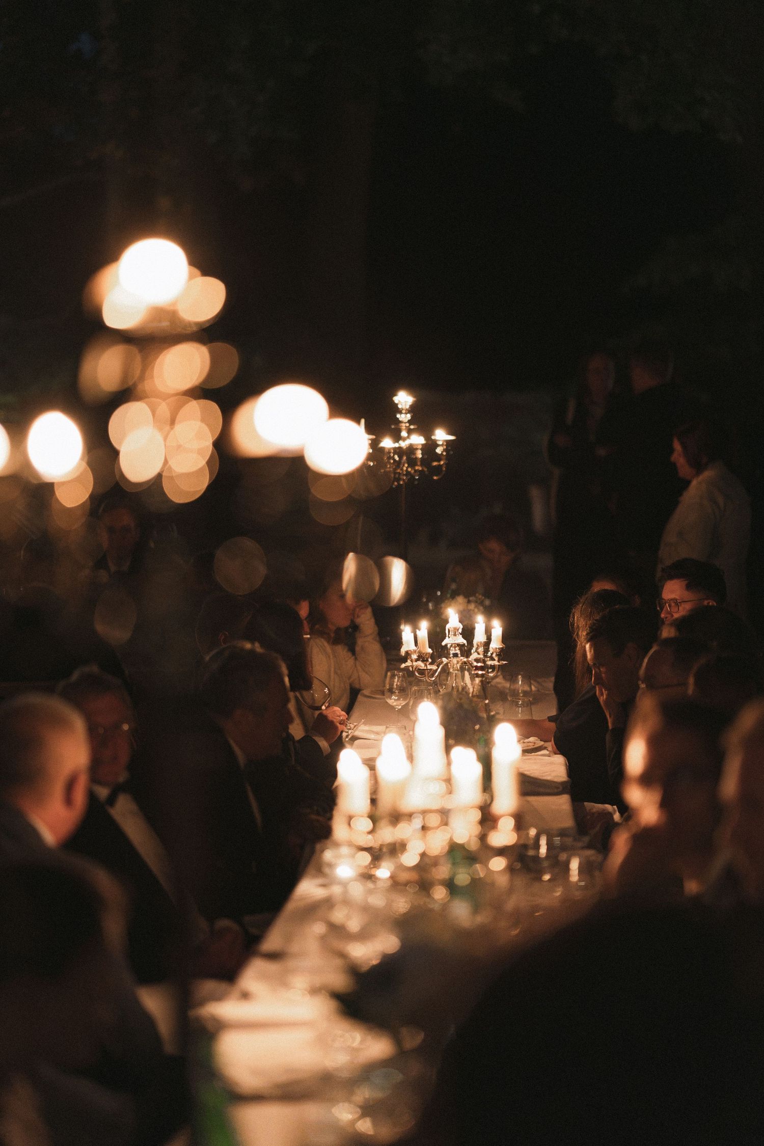 Guests in formal attire dine at a long candlelit table with candelabras and warm glowing lights in a dark, intimate evening setting.