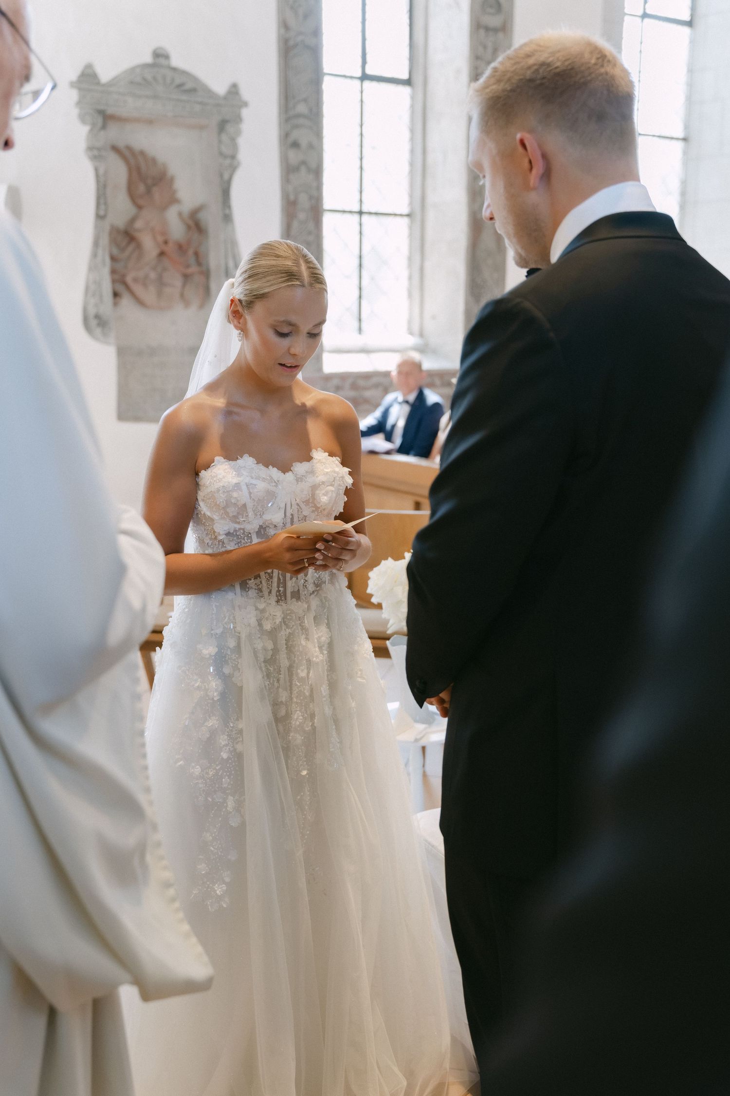 Bride in a strapless floral lace wedding gown holds her vows while facing the groom in a dark suit during a church ceremony.
