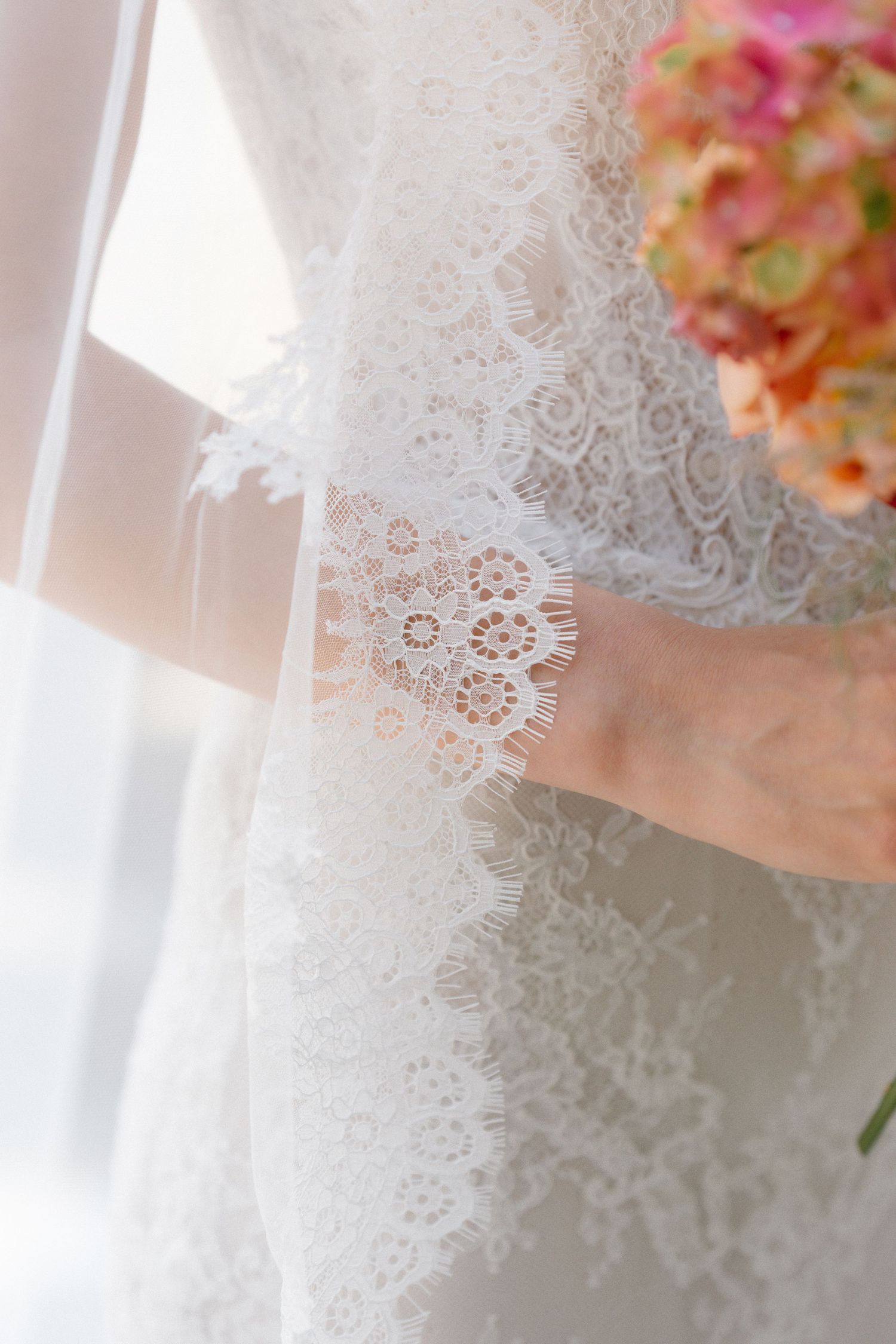 Close-up of a bride in a white lace wedding dress holding a colorful pink and orange floral bouquet.