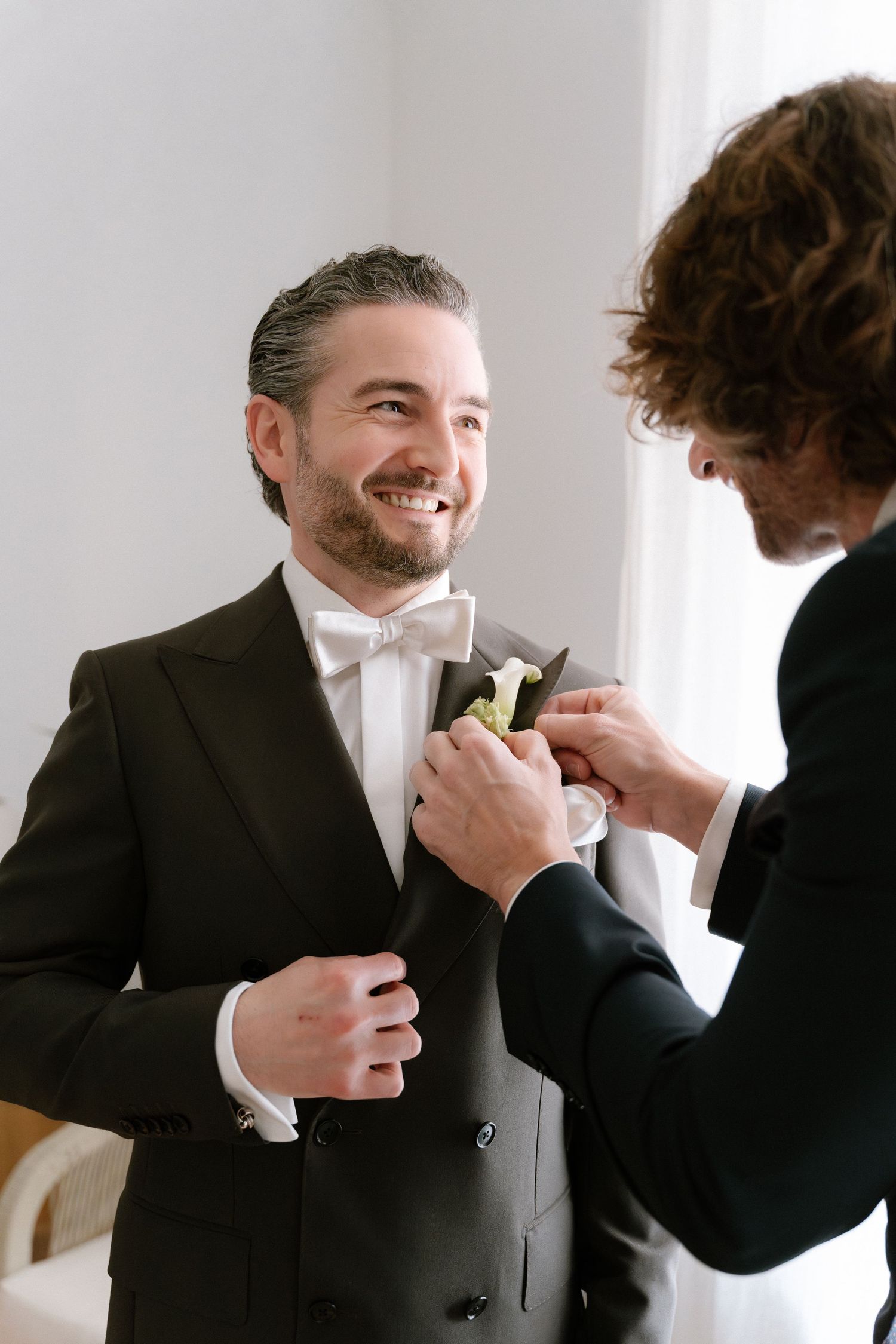 Smiling groom in black tuxedo with bow tie gets boutonniere pinned by best man during wedding preparations.