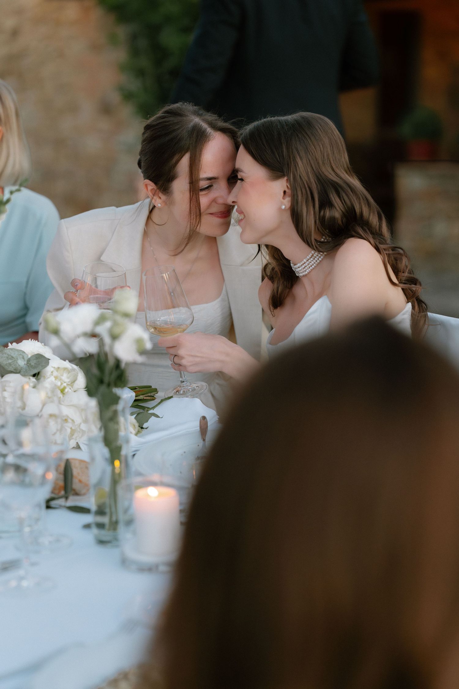 Two brides share an intimate moment at their wedding reception table, holding champagne glasses among white floral arrangements.
