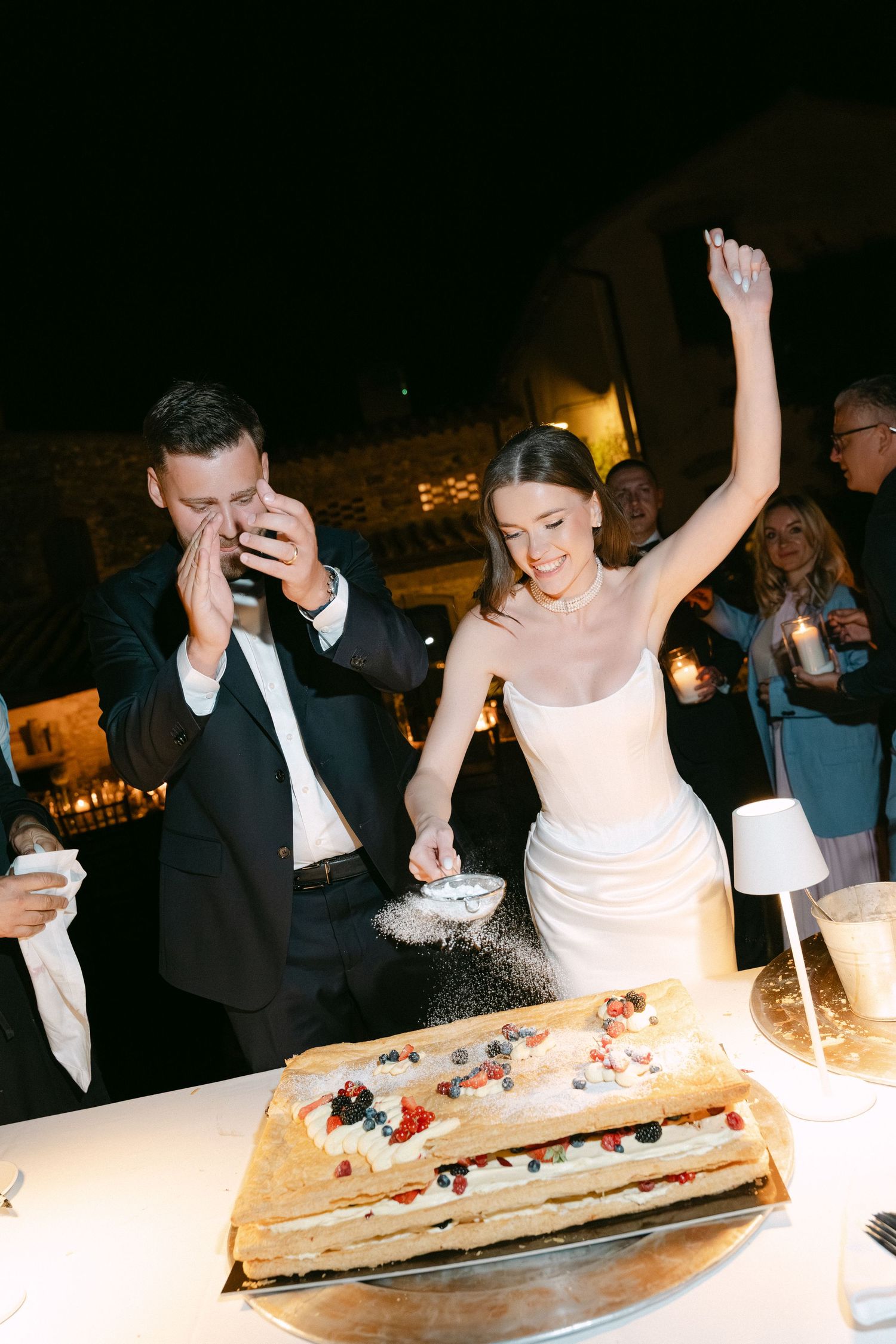 Bride and groom cutting a layered wedding cake topped with fresh berries, bride raising her arm in celebration at evening reception.