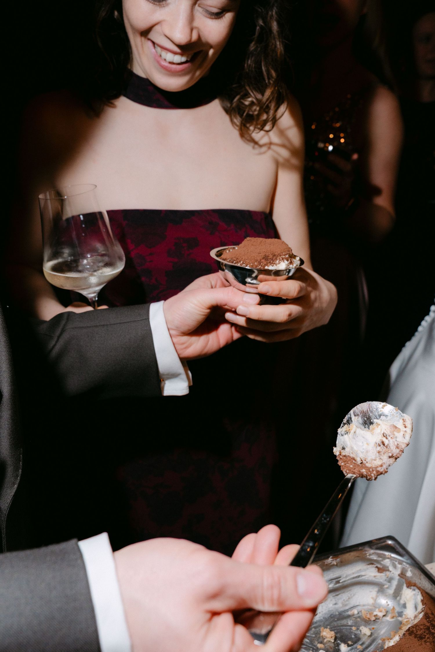 A smiling woman in elegant attire holds a dessert and wine glass at a formal event.