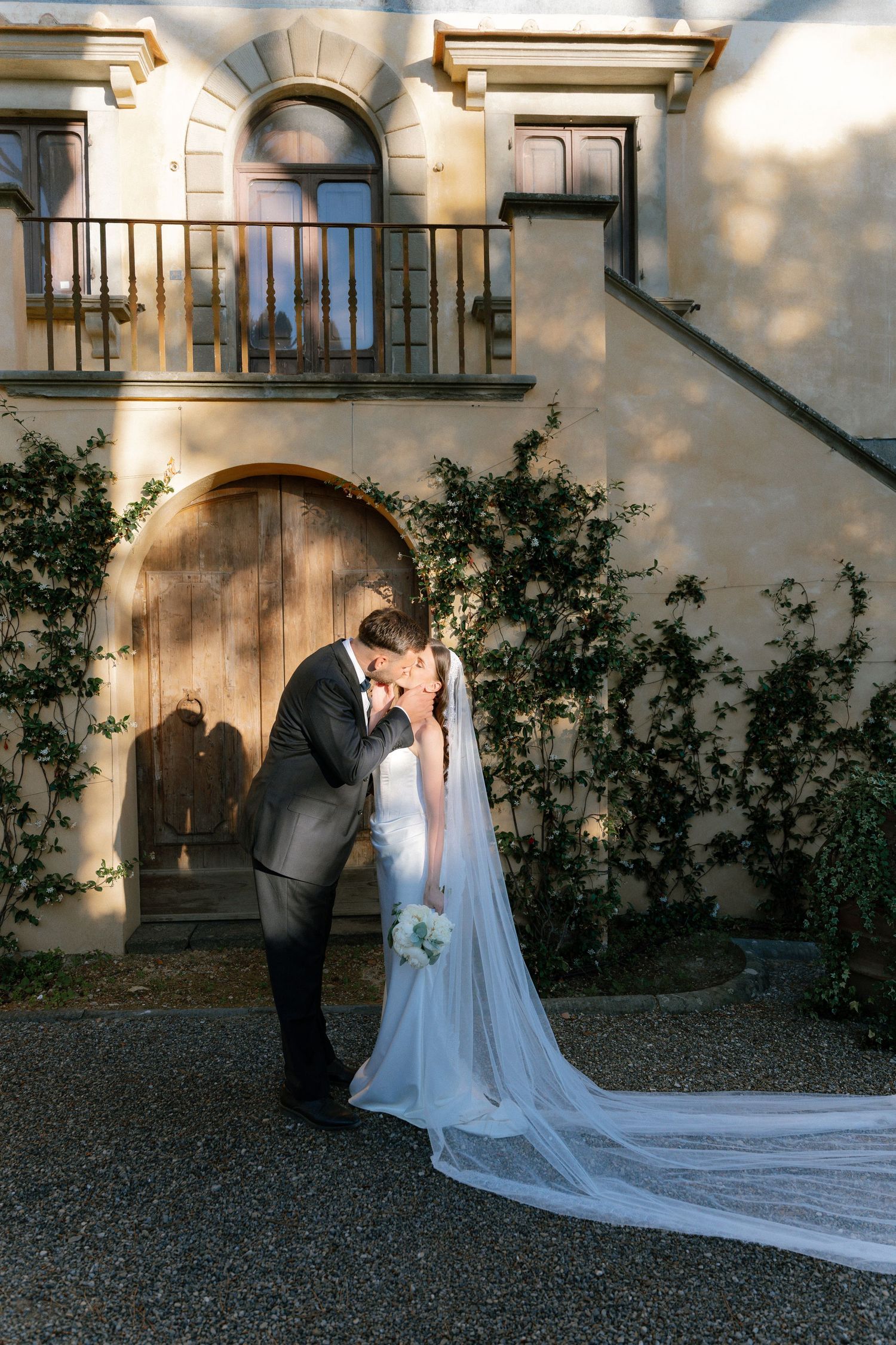 Bride and groom share a romantic kiss in front of a rustic stone building with ivy, bride wearing a long flowing veil.
