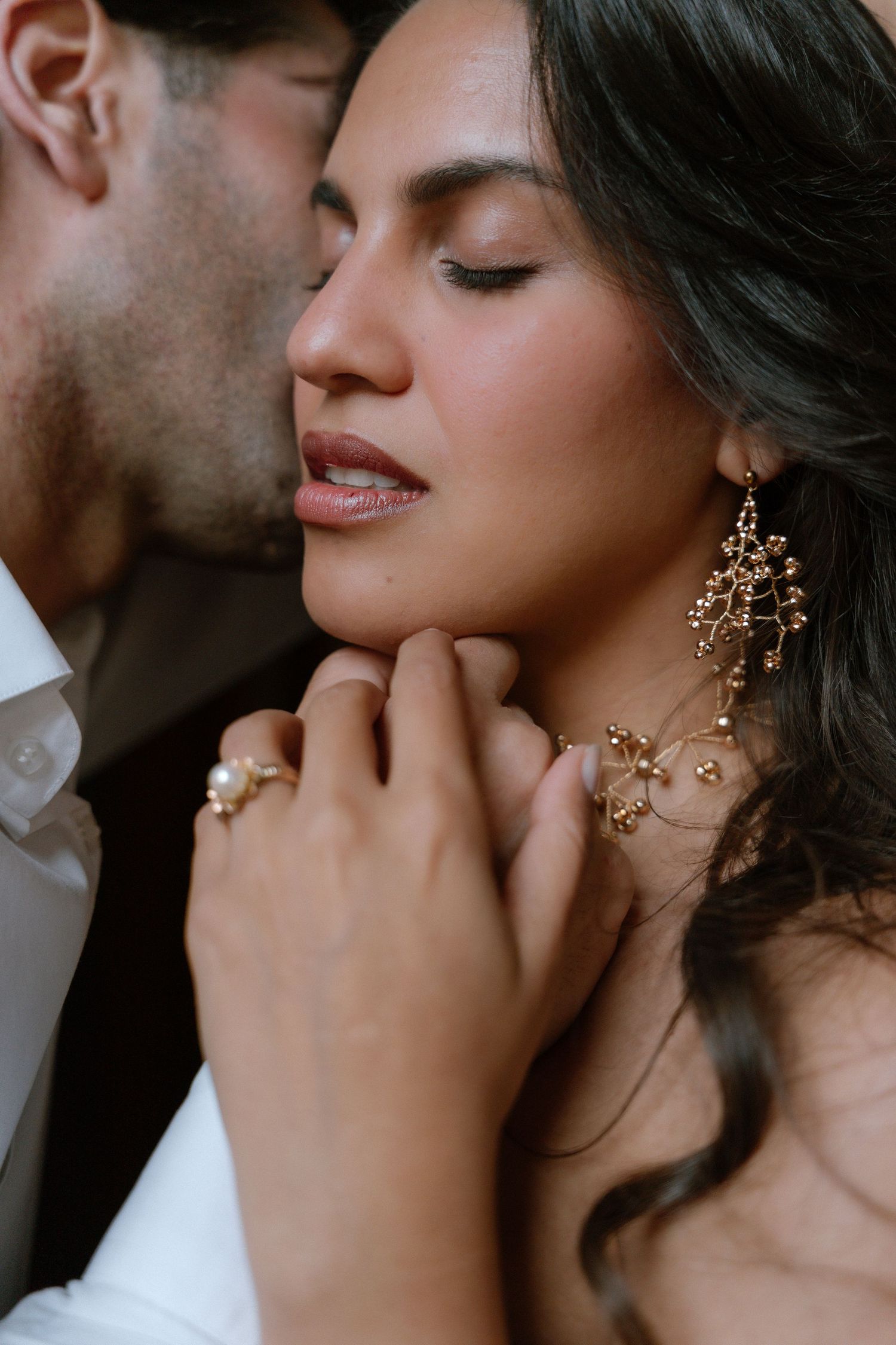 Romantic close-up of a bride with gold chandelier earrings and pearl ring as groom kisses her cheek.
