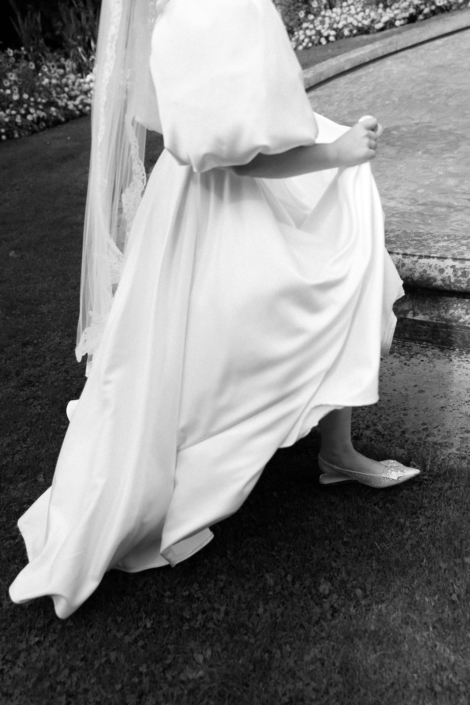 Black and white photo of a bride in a flowing white wedding gown and veil, lifting her dress while walking on wet pavement.
