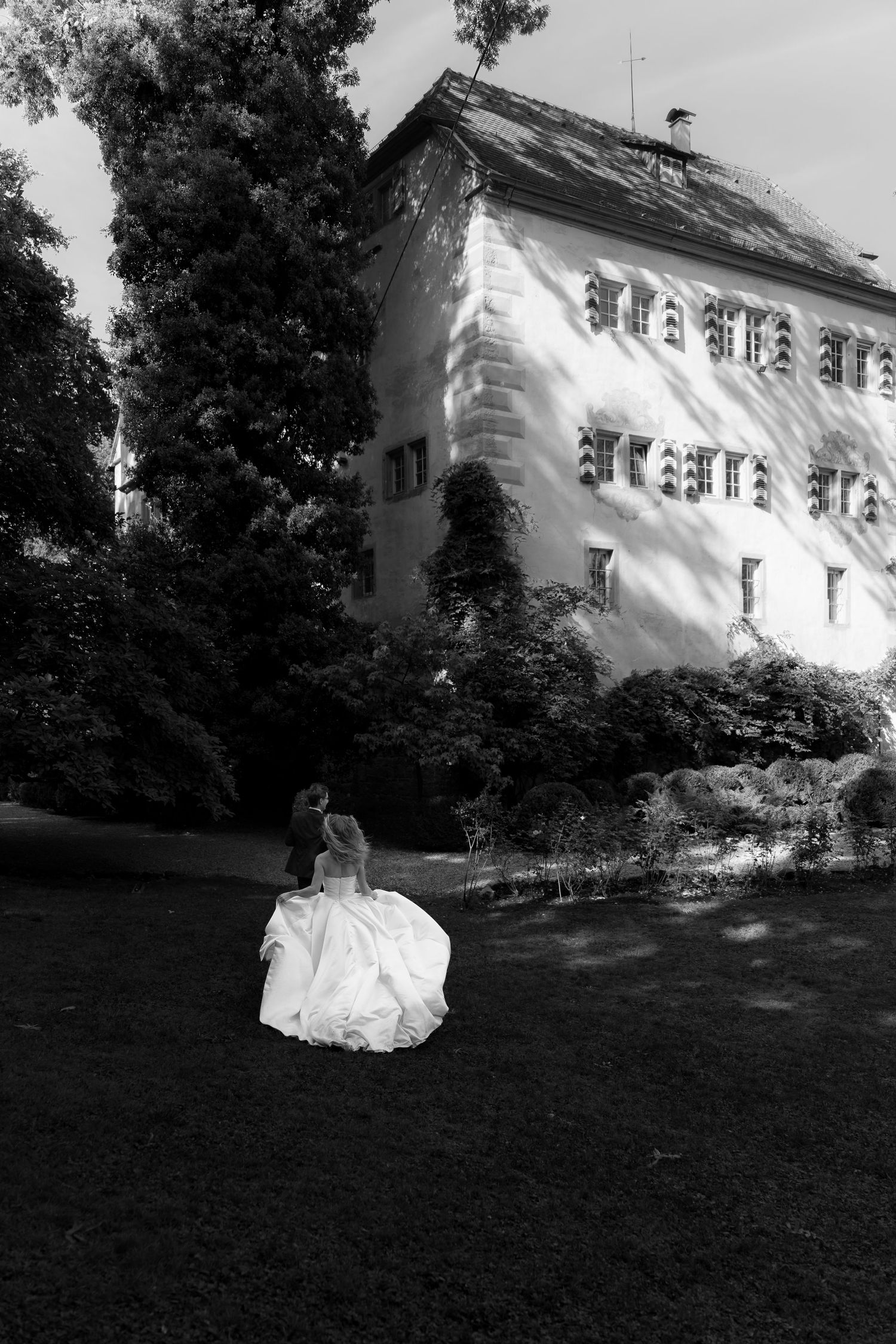 Black and white photo of a bride in a flowing gown walking across a lawn toward a historic manor house surrounded by tall trees.