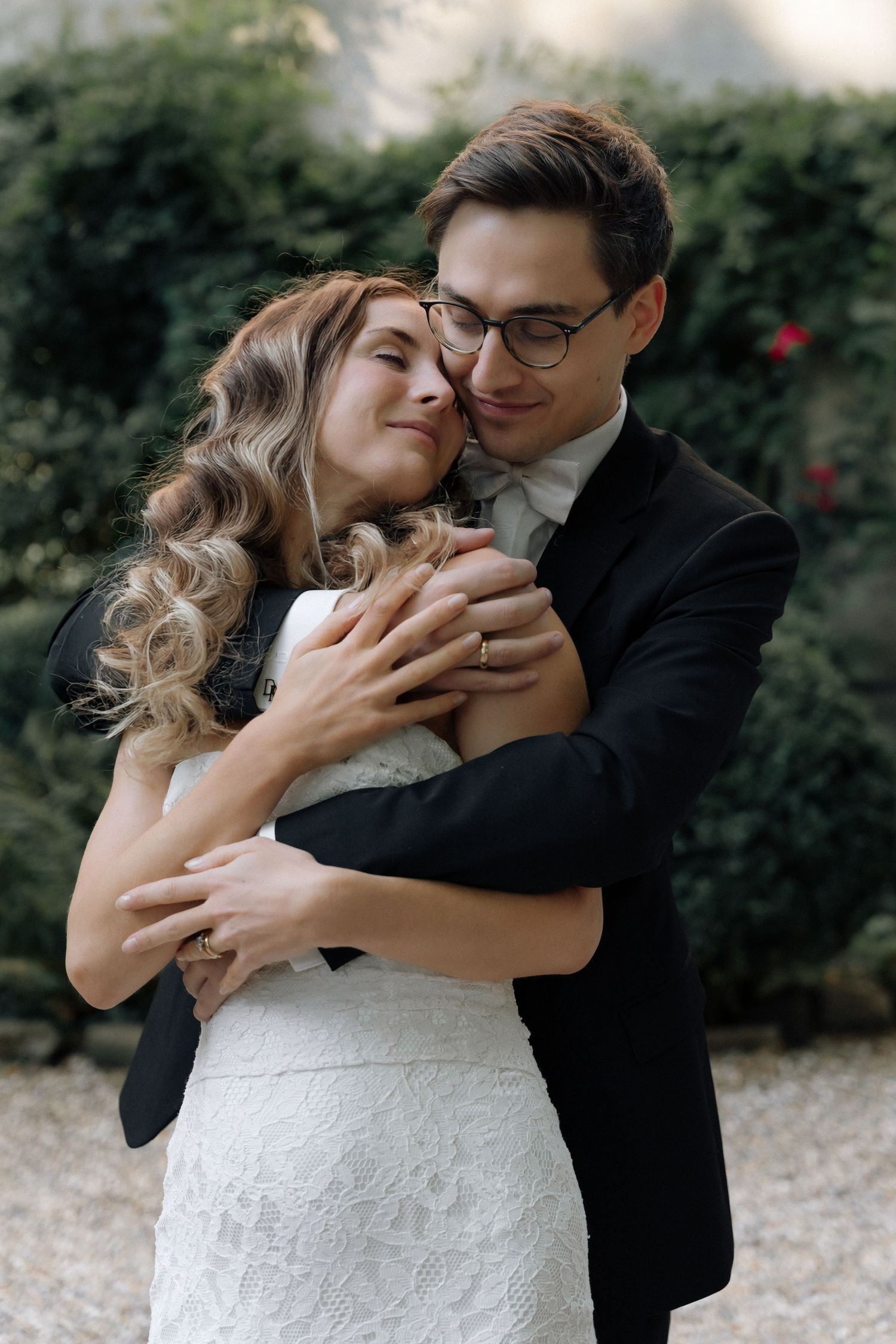 Smiling bride in a lace wedding dress embraced from behind by a groom in a black suit and glasses in a leafy garden.