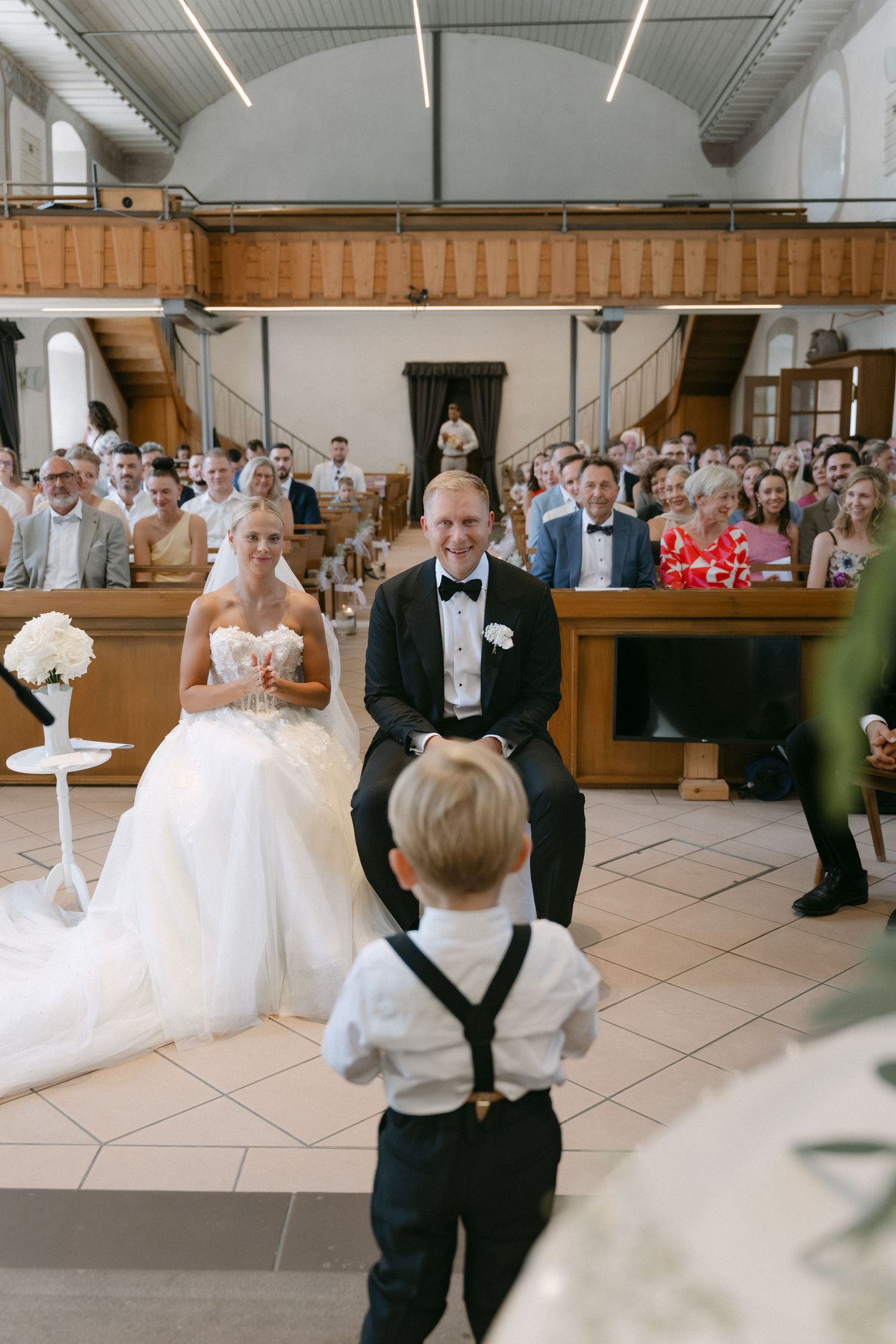 Smiling bride and groom sit at the front of a church as a ring bearer in suspenders faces them and wedding guests look on.