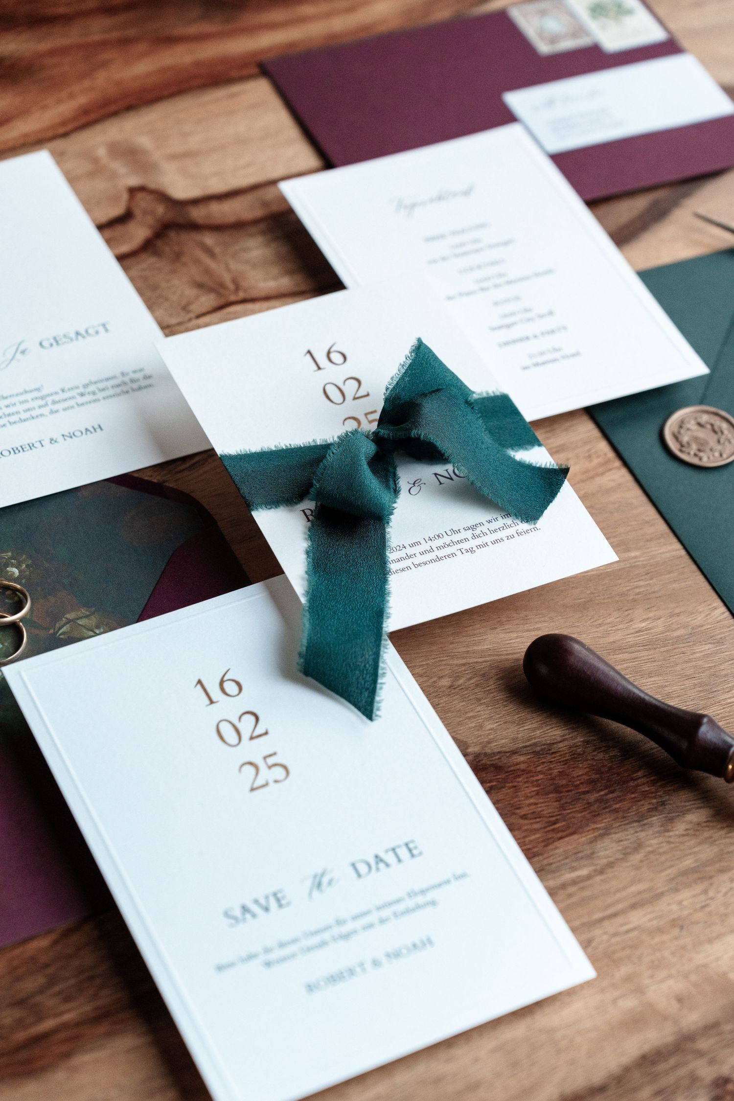 Elegant wedding invitation suite featuring a teal ribbon bow, white cards with date details, and burgundy envelopes on wood surface.