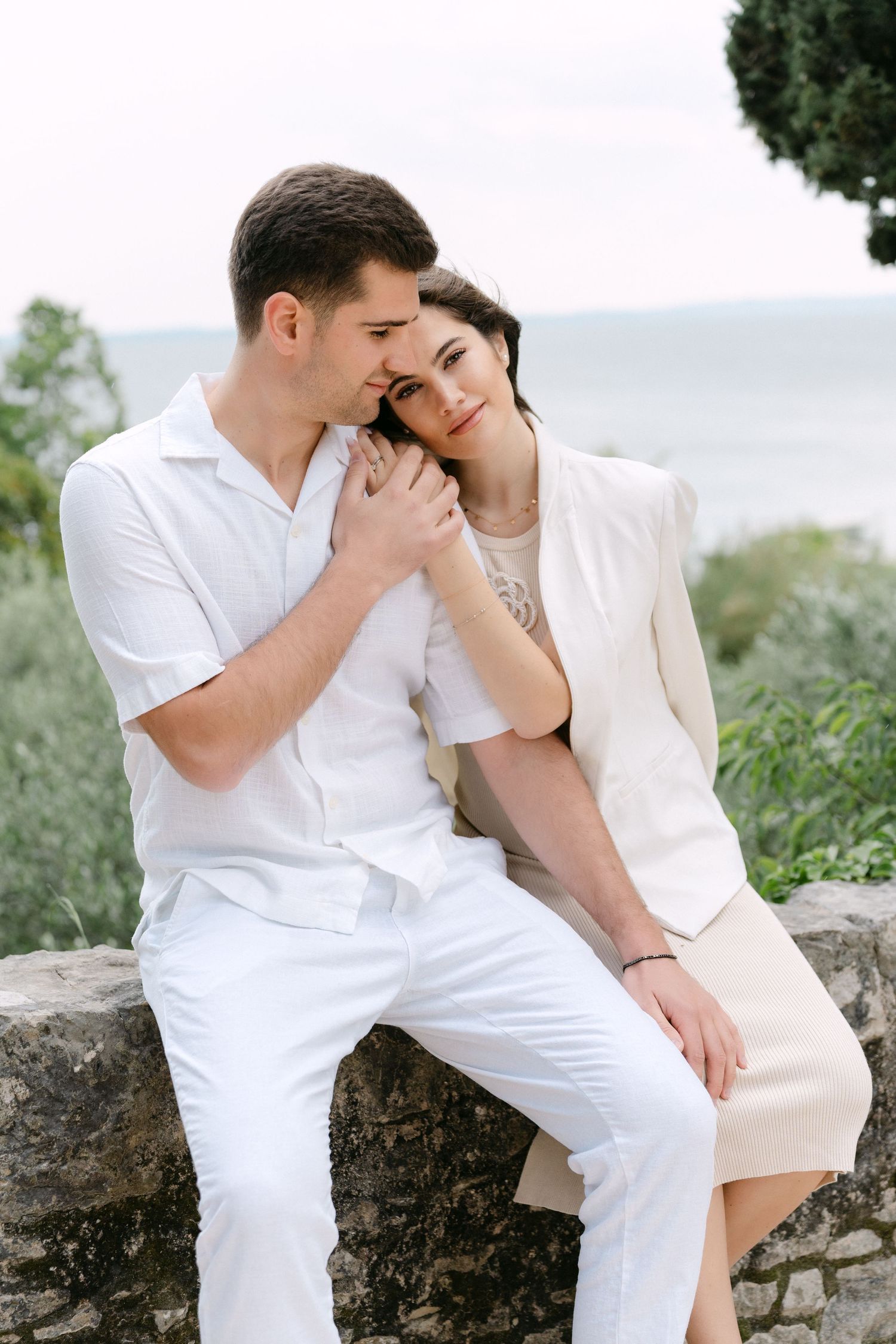 A romantic couple dressed in white outfits share an intimate moment while sitting on a stone wall with nature in background.