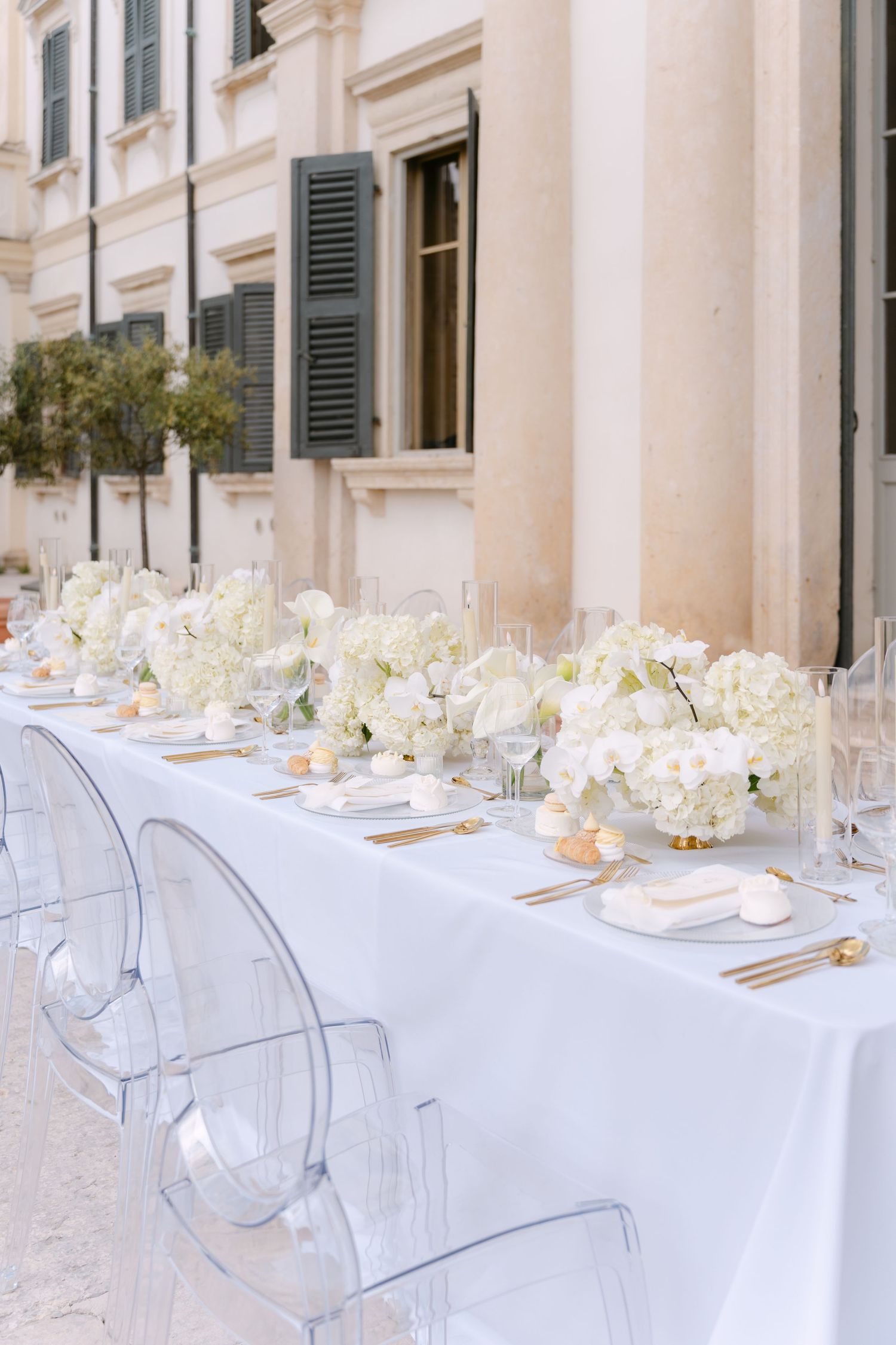 Elegant outdoor table setting with white linens, ghost chairs and white floral centerpieces along a classic villa facade. Villa Mosconi