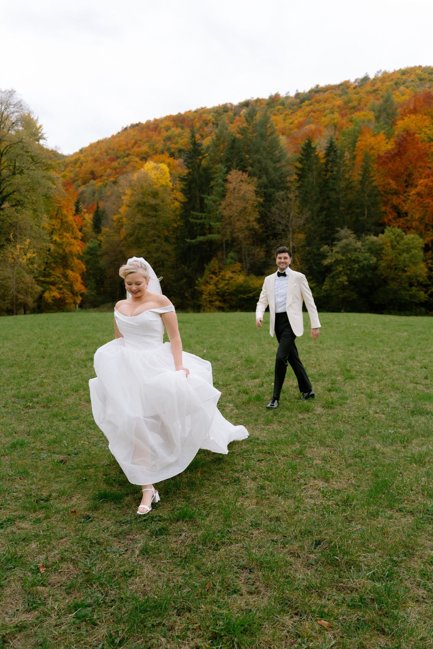 A couple in wedding attire runs playfully across a grassy field with vibrant autumn foliage in the background.