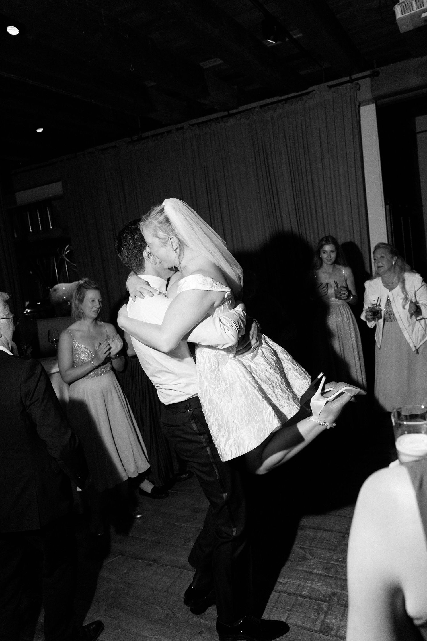A couple in wedding attire share a passionate dance dip at their reception while guests look on in the background.
