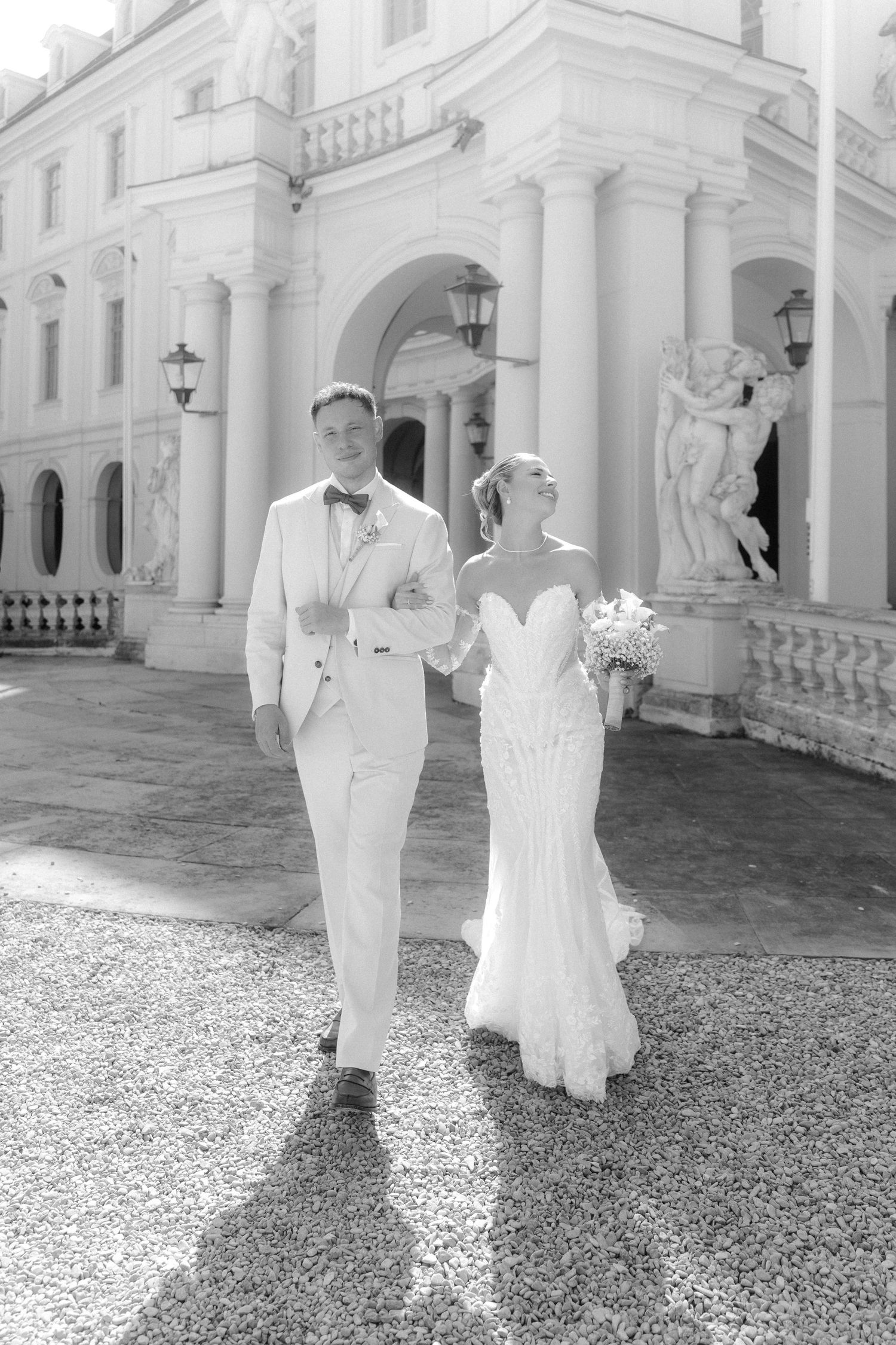 Wedding couple in white attire walking along a grand white columned building exterior.