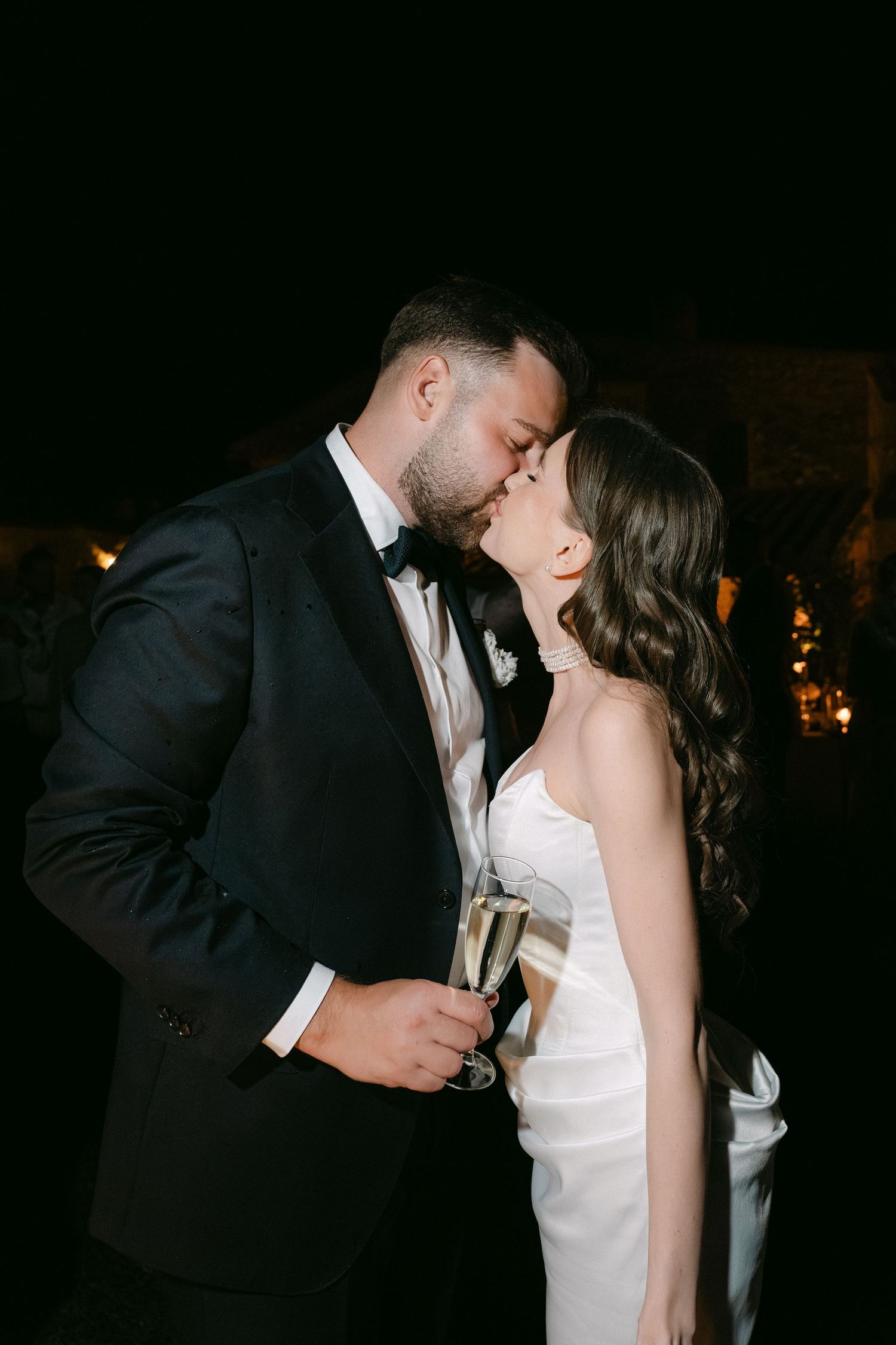 Newlyweds share a romantic kiss at night, groom in black tuxedo holding champagne, bride in white strapless gown.