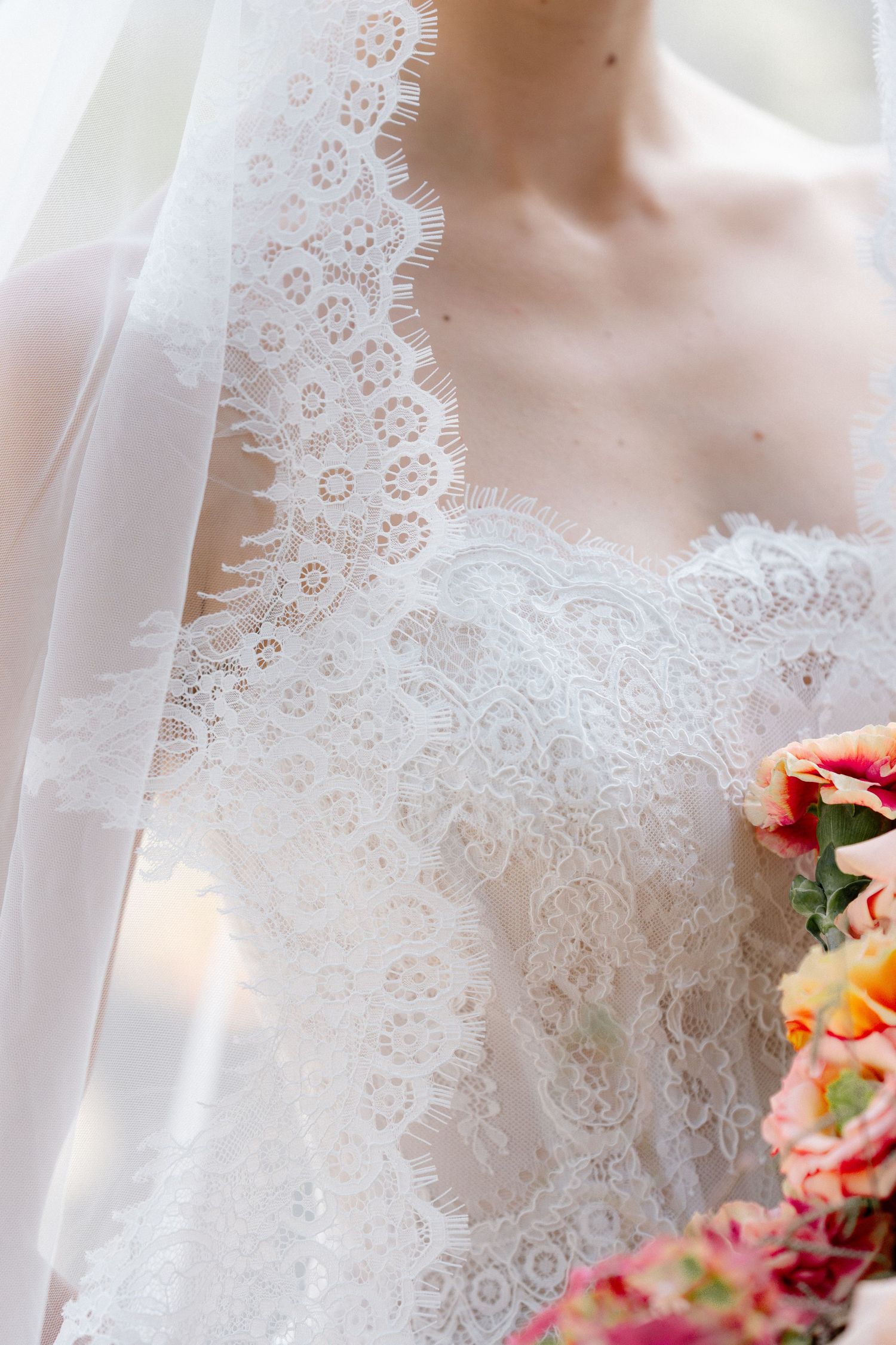 Bride in elegant white lace wedding gown holding colorful bridal bouquet with delicate veil detail.