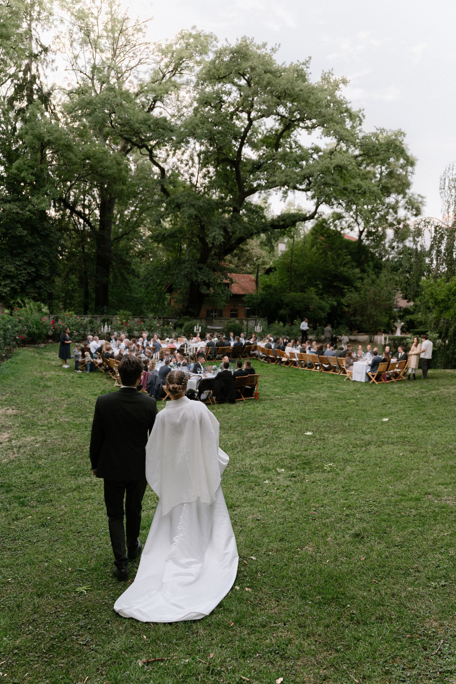 Bride in a white gown and groom in a dark suit walk across a grassy lawn toward guests seated at an outdoor garden wedding ceremony.