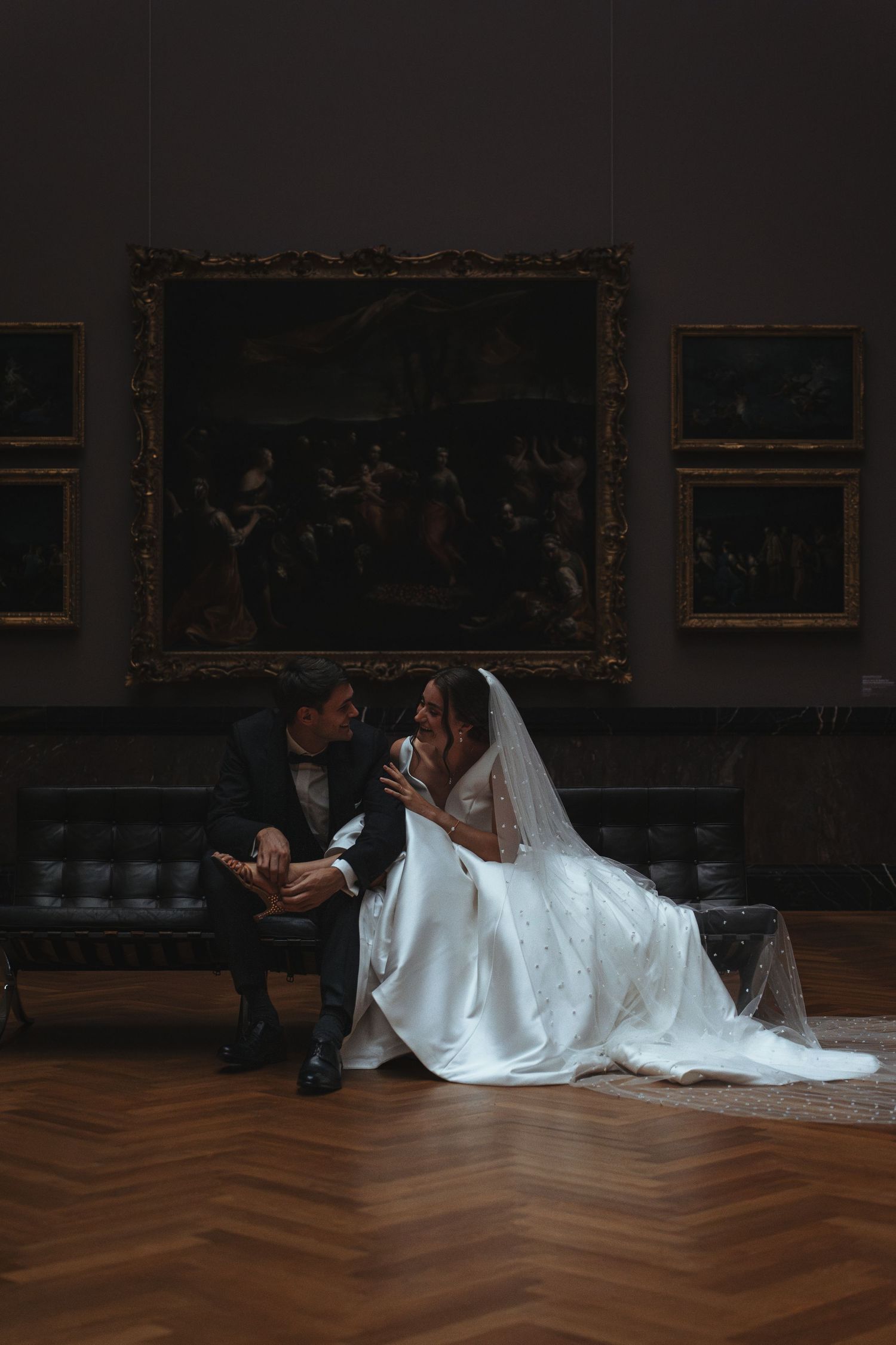 A romantic wedding photo captured in a museum gallery with ornate paintings and herringbone floors.