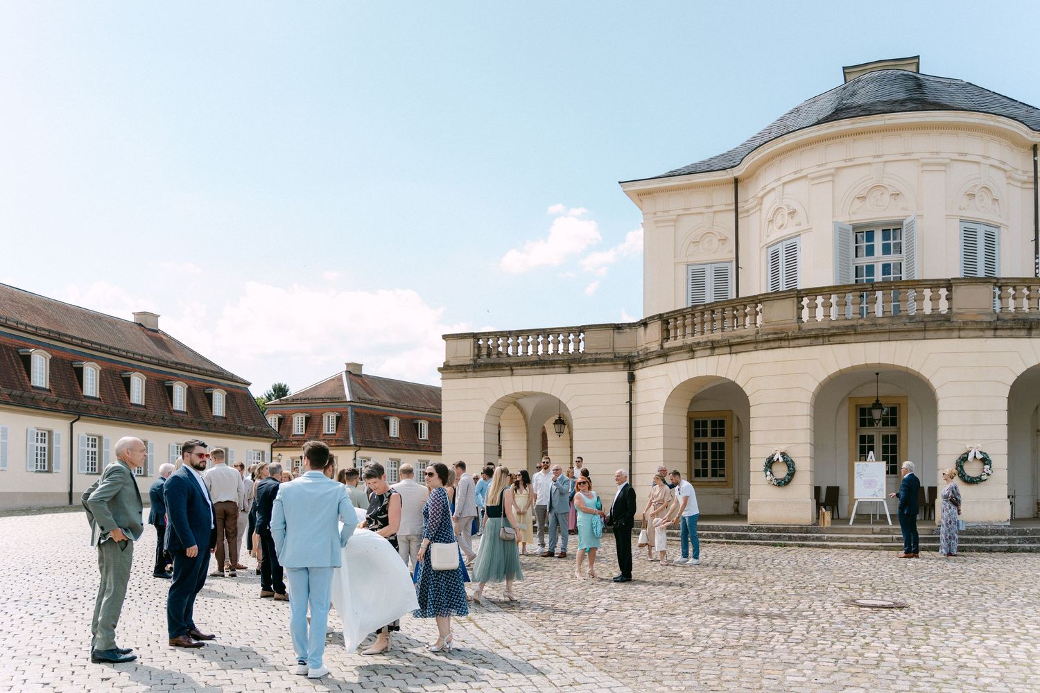 Panoramic view of tourists exploring a historic palace courtyard with stone buildings and classical architecture. Schloss Solitude