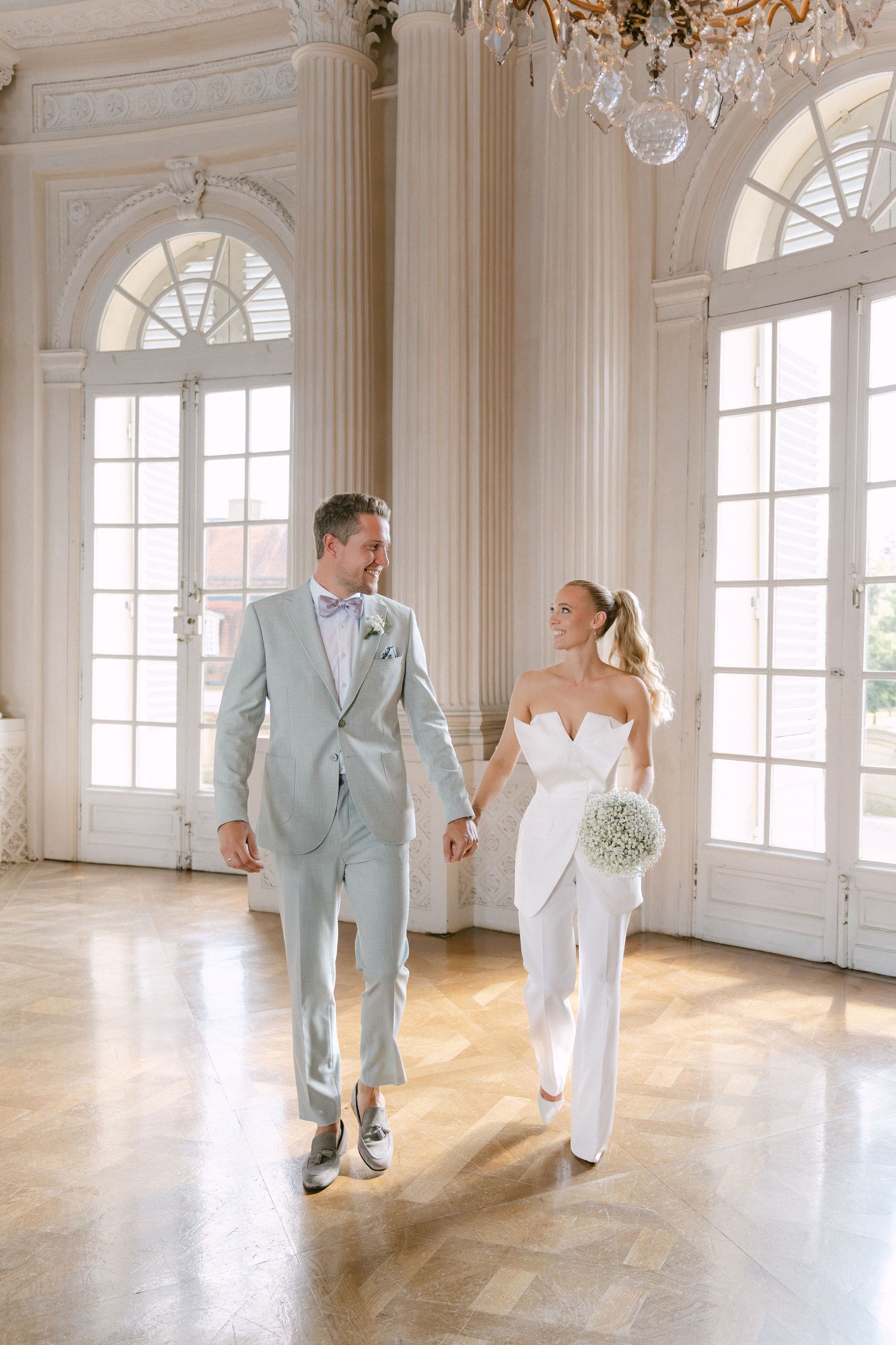 A couple in wedding attire dances playfully across an elegant white ballroom with arched windows and natural light. Schloss Solitude