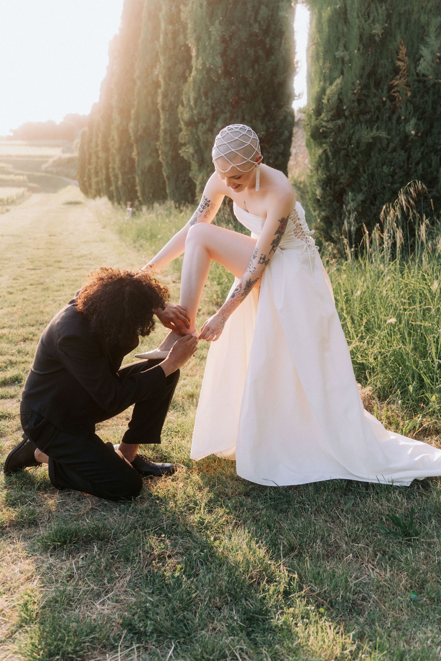 A person in a white wedding dress pets a black dog in a sunlit outdoor setting.