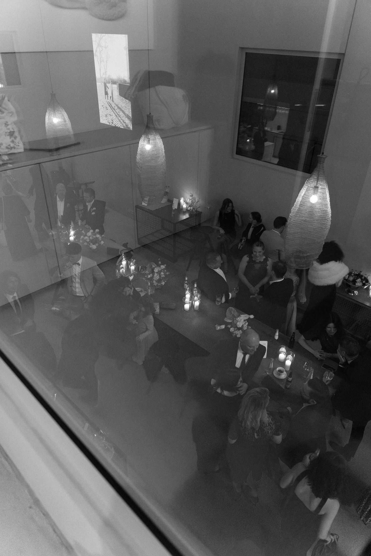 Black and white aerial view of elegant indoor party with hanging lanterns and guests socializing around candlelit tables.