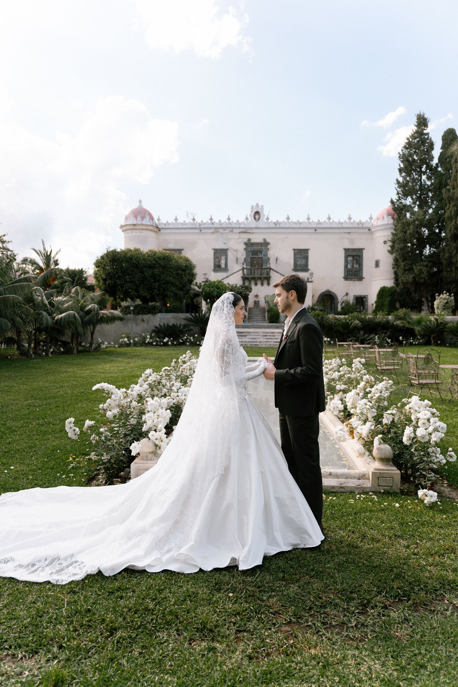 Bride in white gown with long veil and groom in black suit stand in lush garden before elegant white mansion.