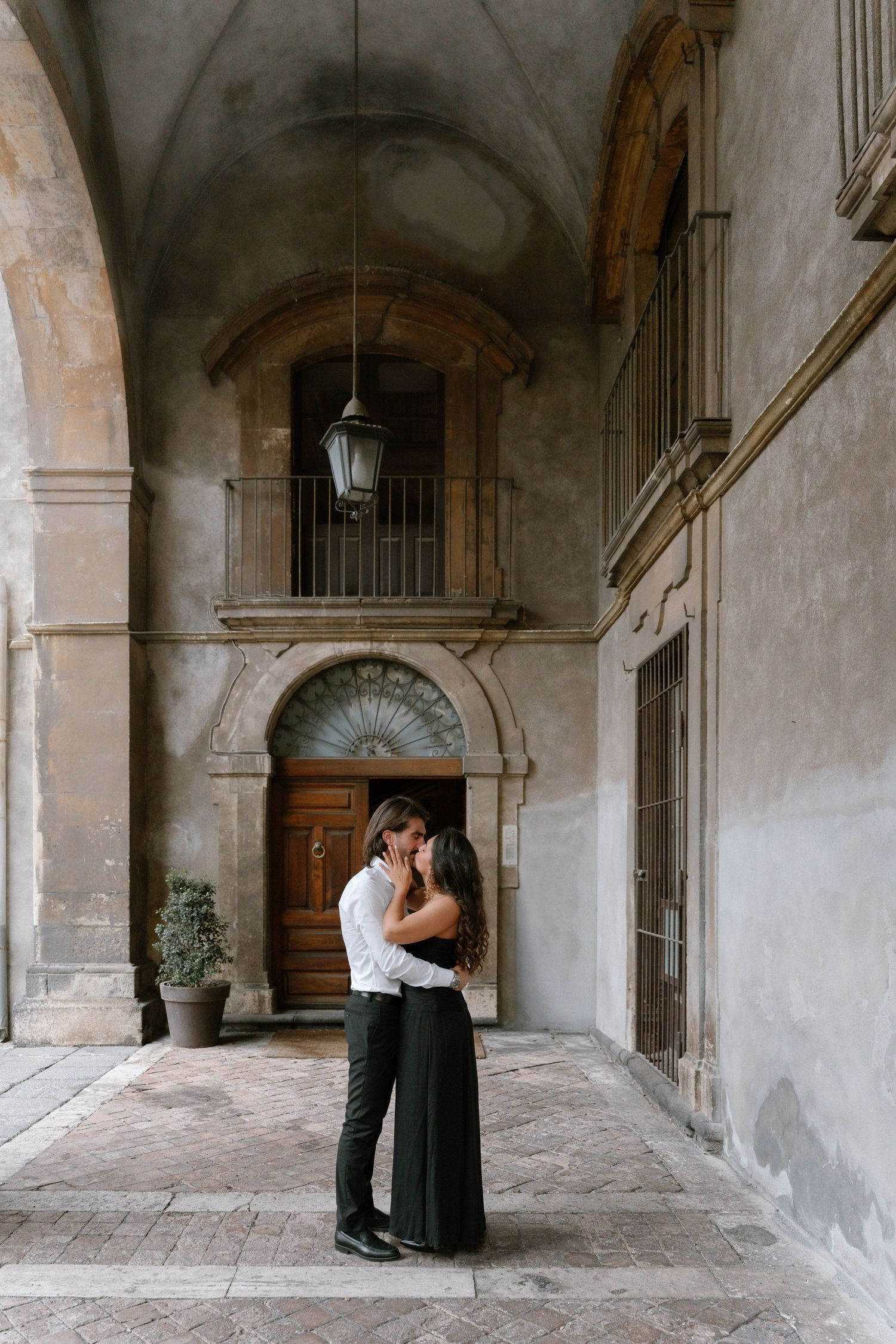 Couple embracing and kissing in an elegant Italian courtyard with arched doorways and vintage lantern.