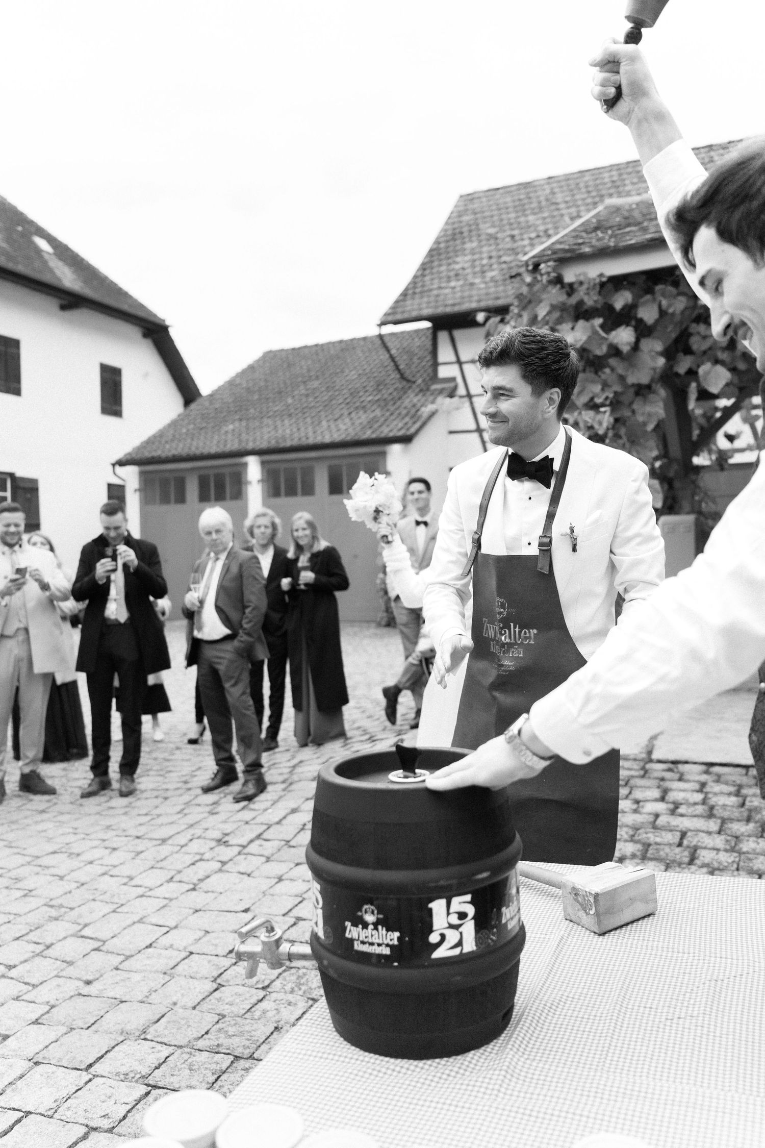 A server in formal attire taps a beer keg at an outdoor event while onlookers gather in the background.
